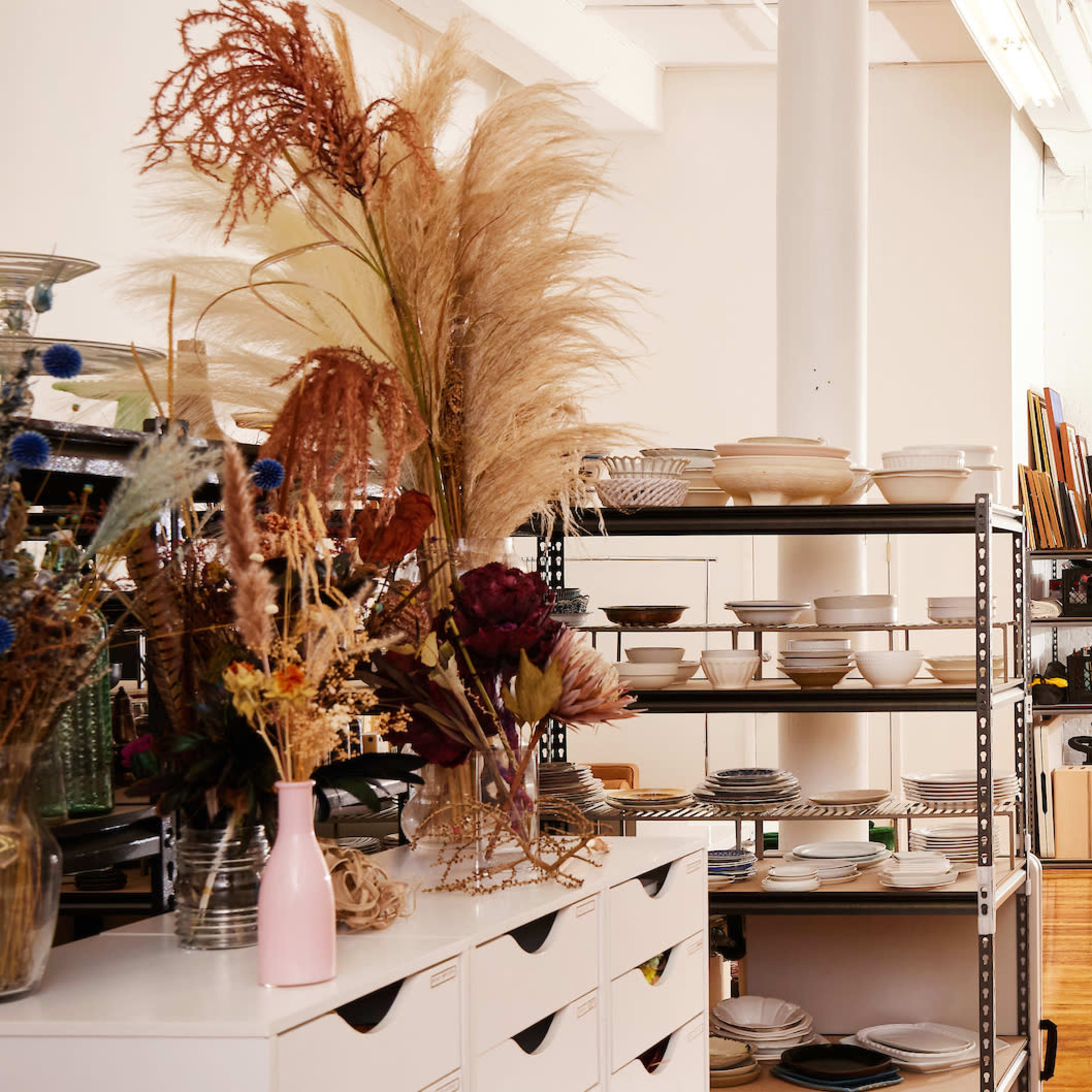 A bright, open space filled with shelves of various home goods, including plates, books, and decorative items, with large windows allowing natural light to enter.