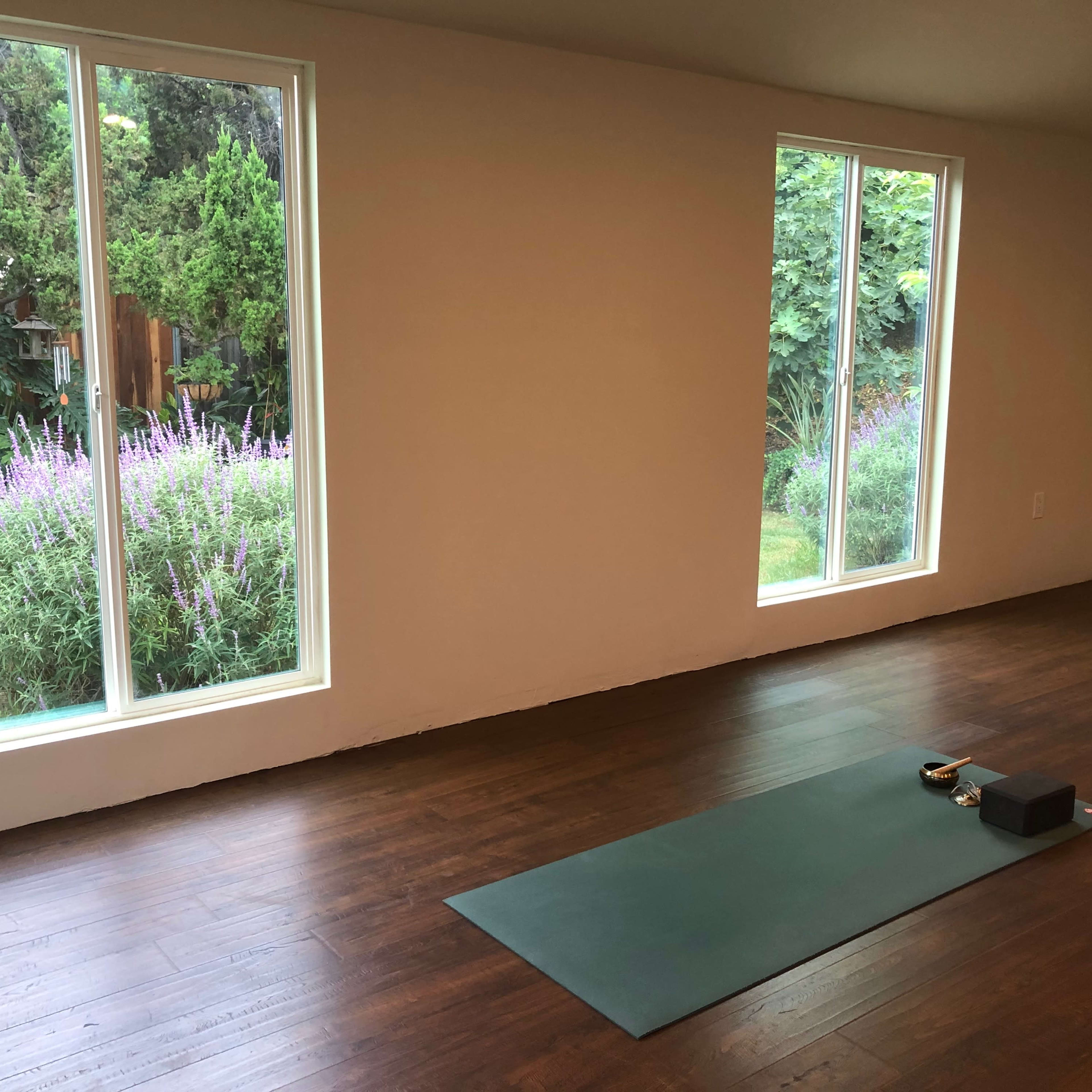 A yoga mat with props is placed on a wooden floor in a room featuring large windows overlooking a garden.