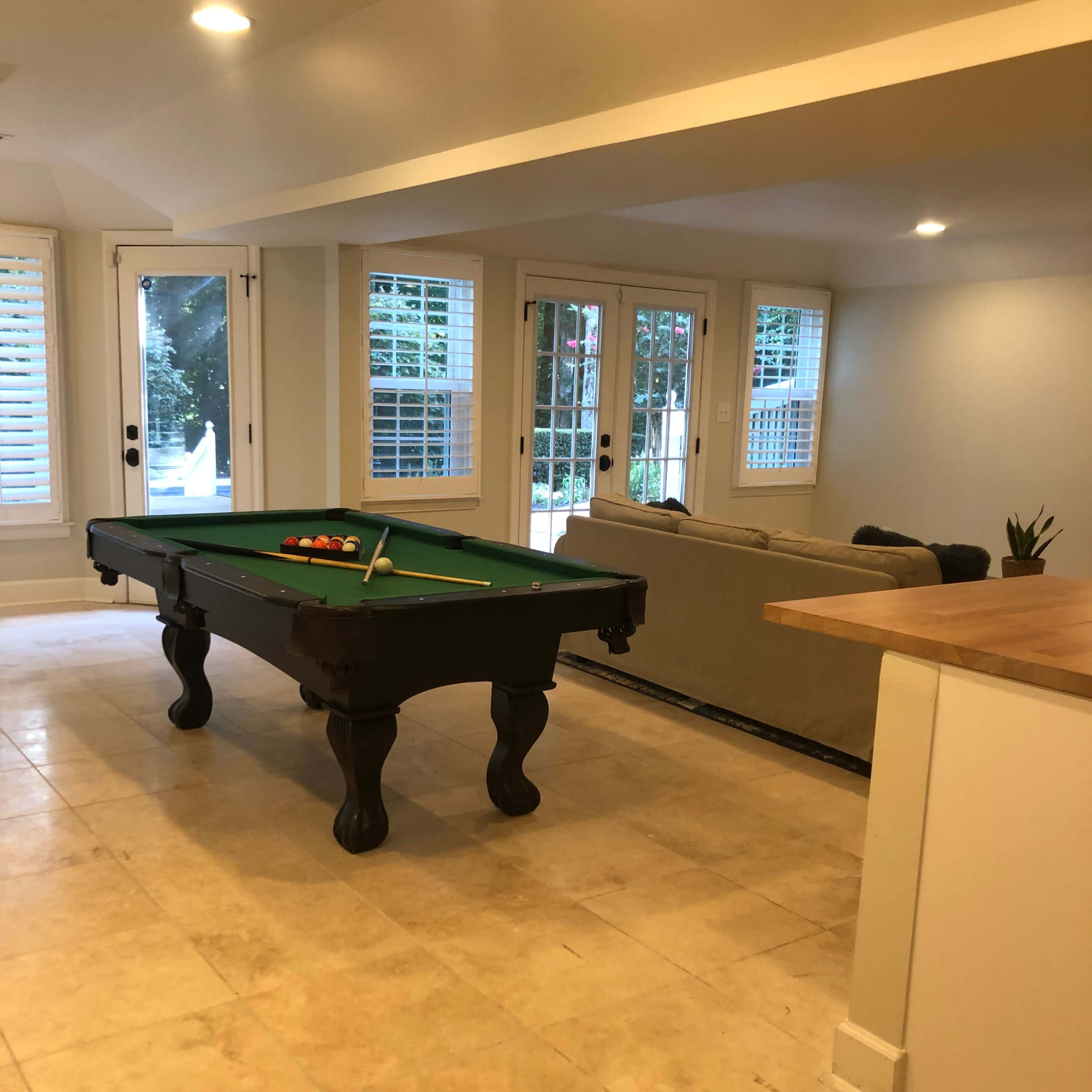 A spacious room featuring a green felt pool table in the center, with a couch and large windows in the background.