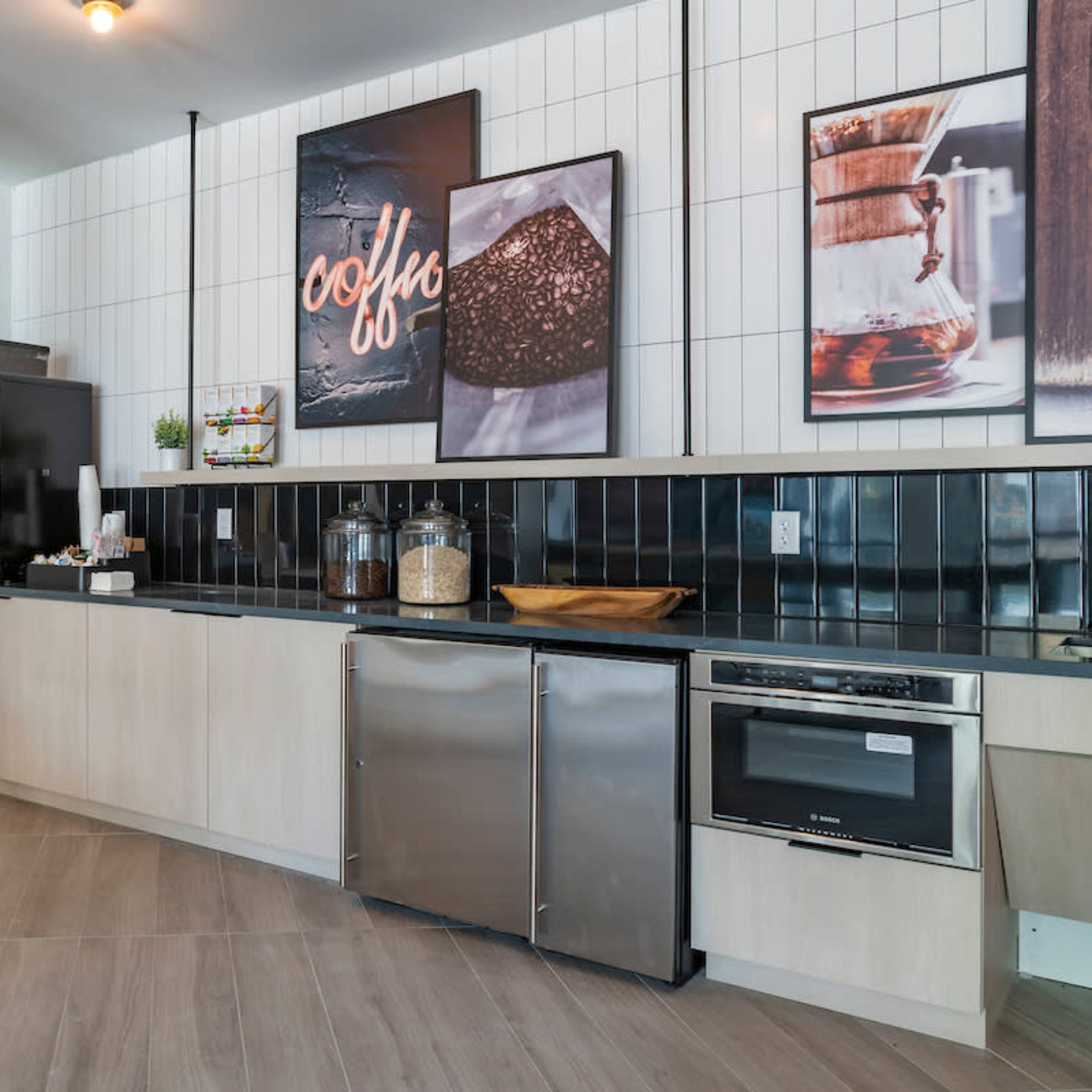 A modern kitchen area featuring a black and white tiled backsplash, wooden shelves with books, and various coffee-related artwork on the walls.