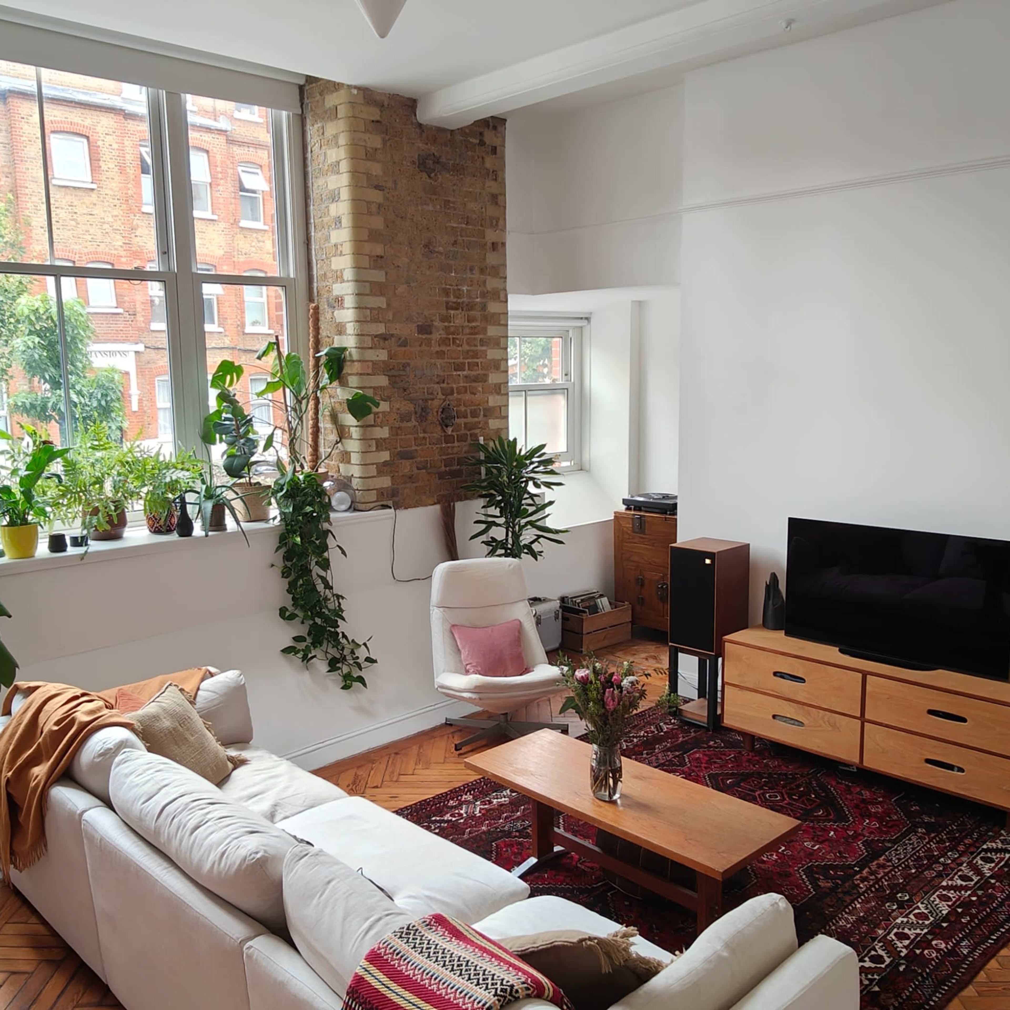 A bright living room with large windows, a white sofa, a wooden coffee table, and various potted plants lining the window sill.
