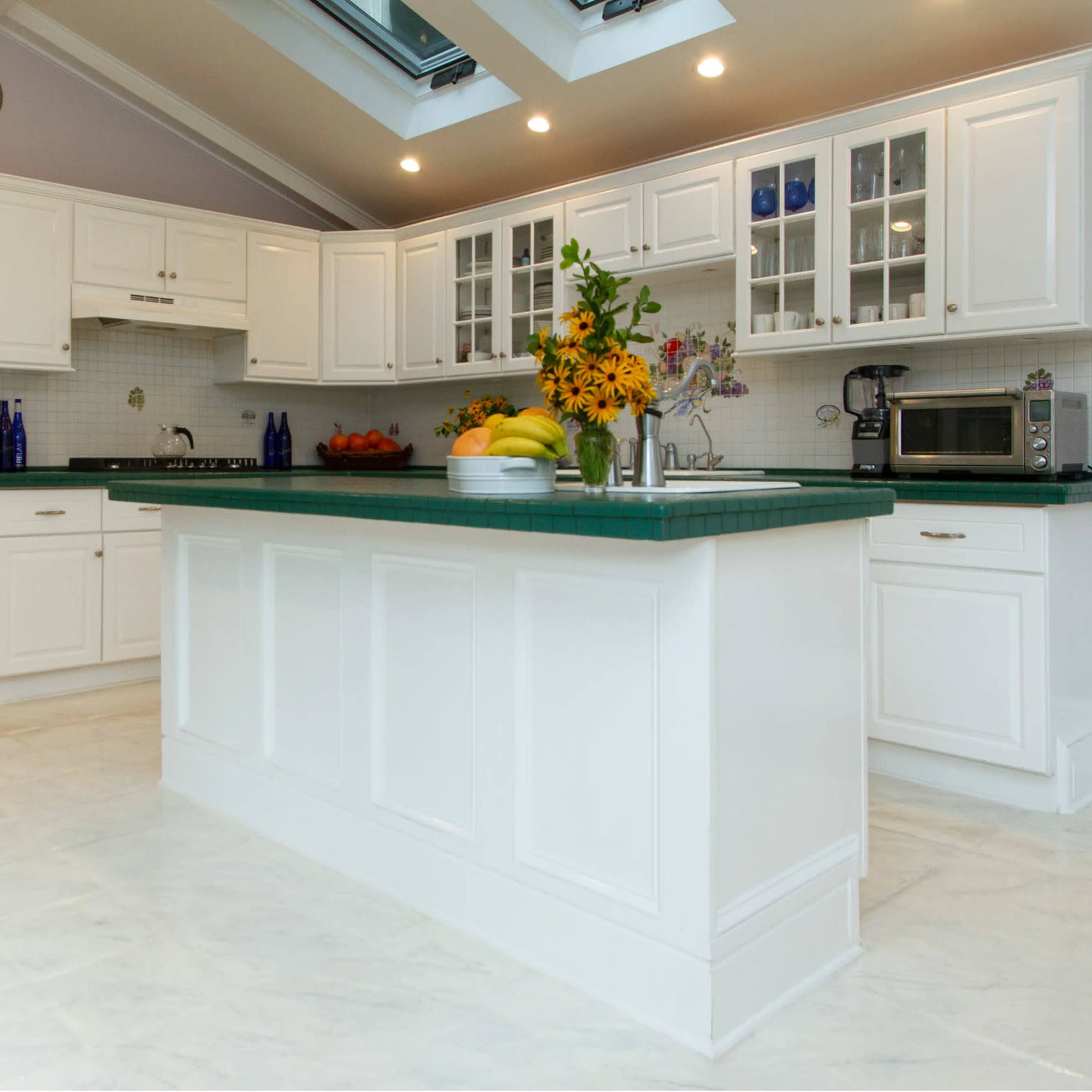 A spacious kitchen with white cabinetry, a green countertop, and a central island featuring a vase of flowers and a bowl of fruit.