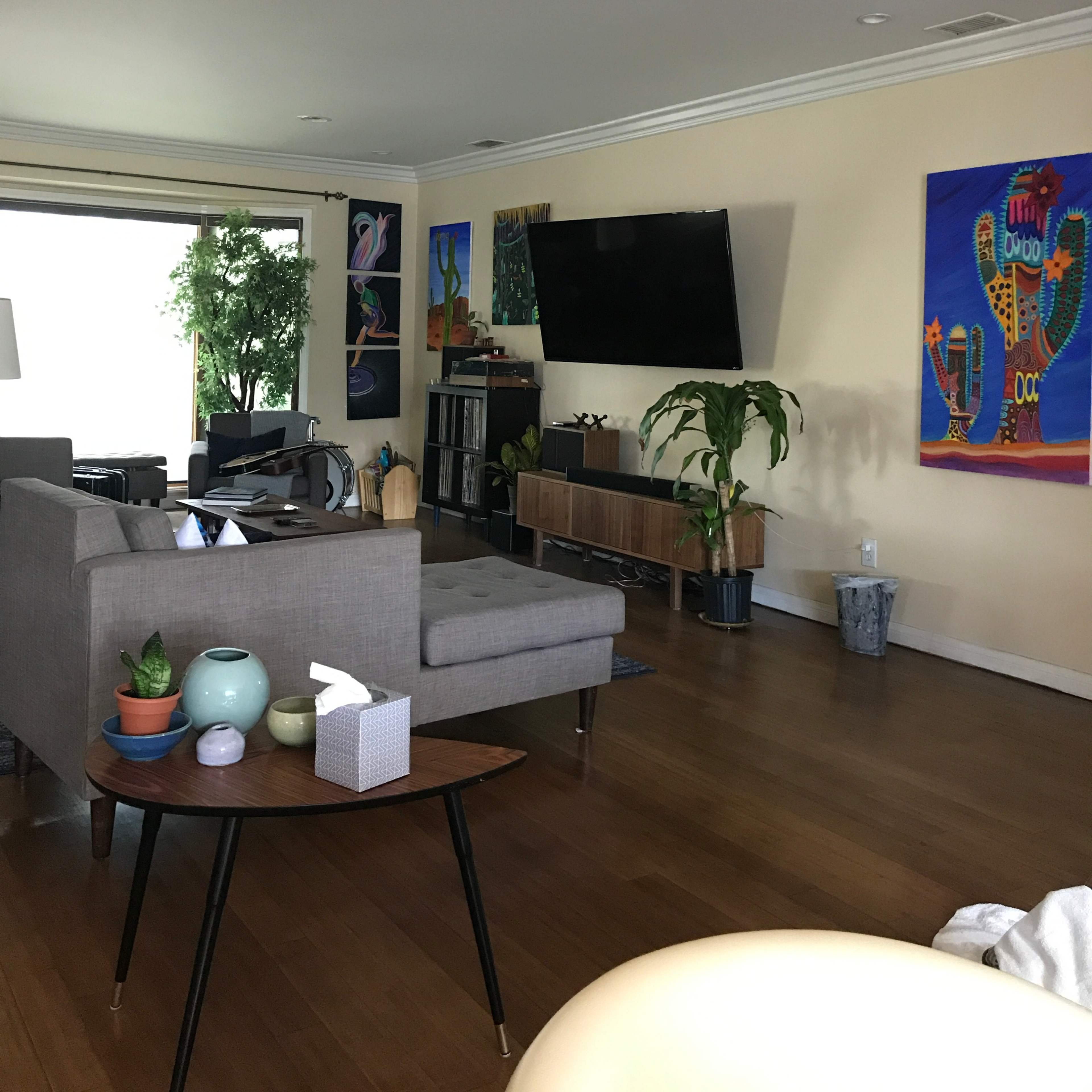 The room features a modern living area with a gray sofa, a wooden coffee table, colorful artwork on the walls, and a large window allowing natural light to enter.