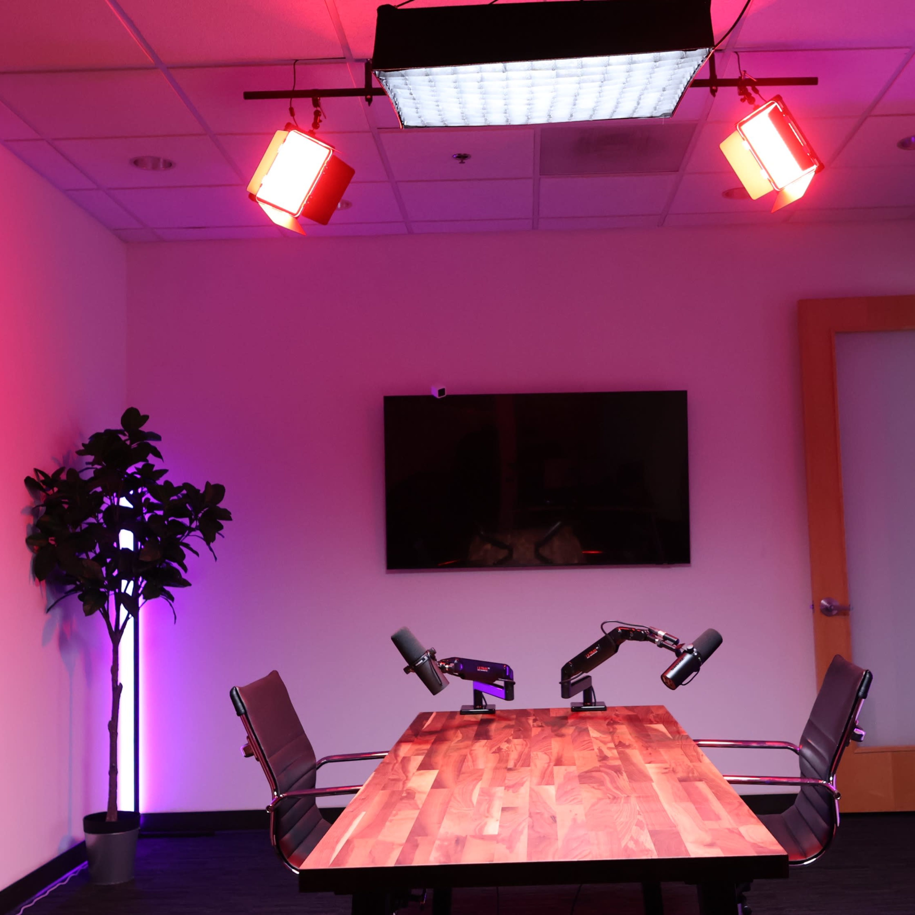 A modern conference room with a wooden table, two microphone arms, a large wall-mounted screen, and vibrant colored lighting.