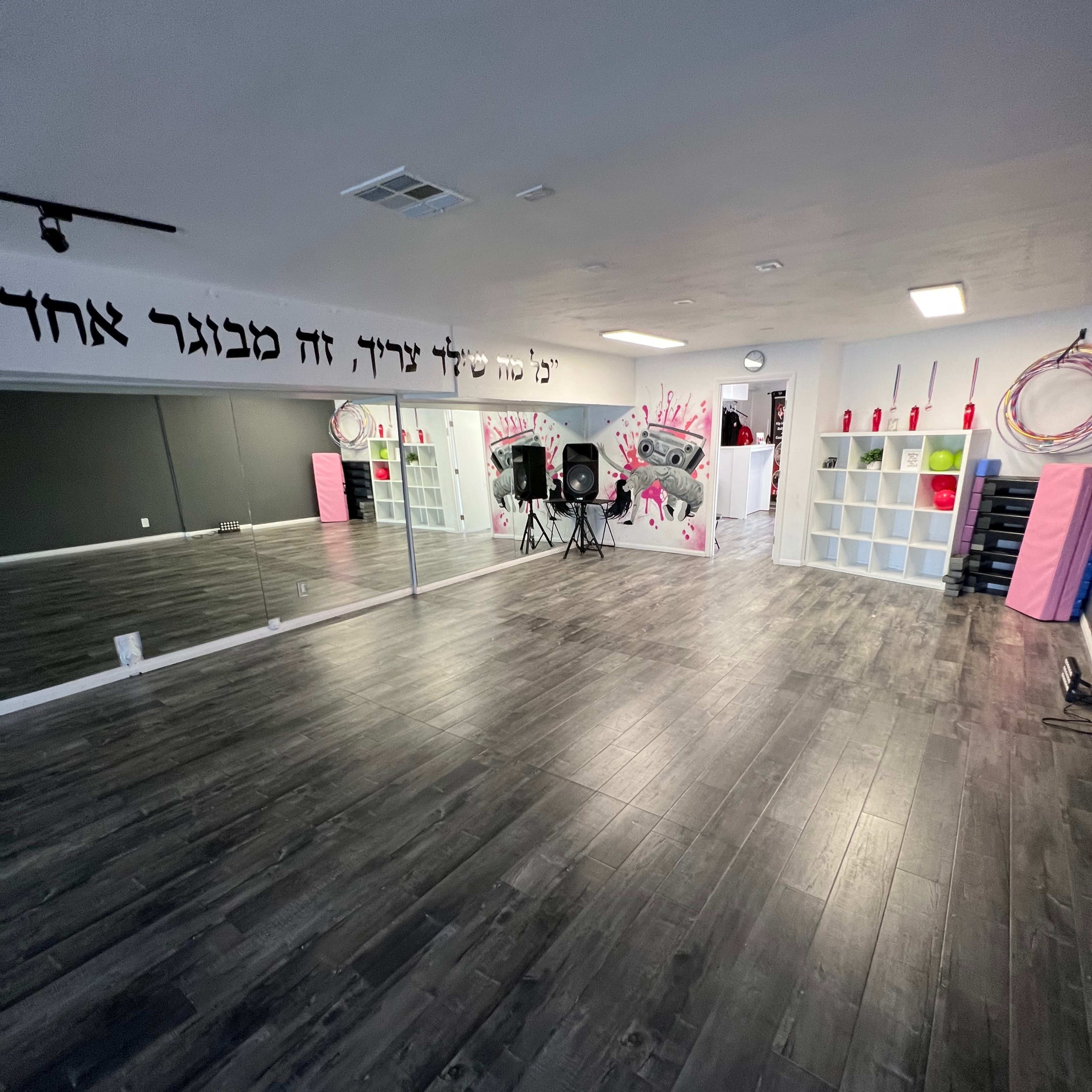 An empty dance studio with a mirrored wall, black flooring, and storage shelves adorned with pink and white accents.