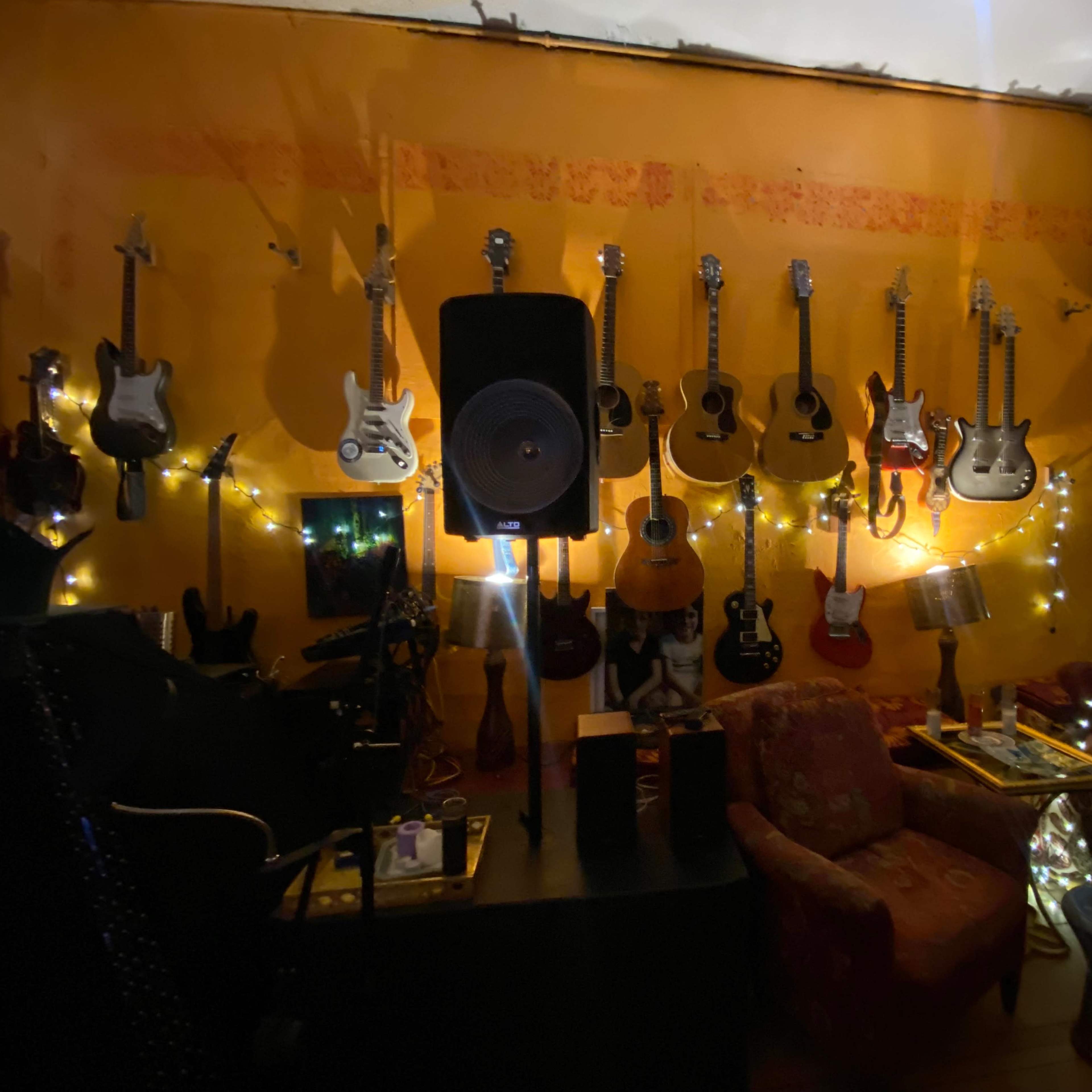 A cozy room with a vibrant orange wall adorned with various guitars, a speaker, and soft lighting from string lights, alongside two upholstered chairs.
