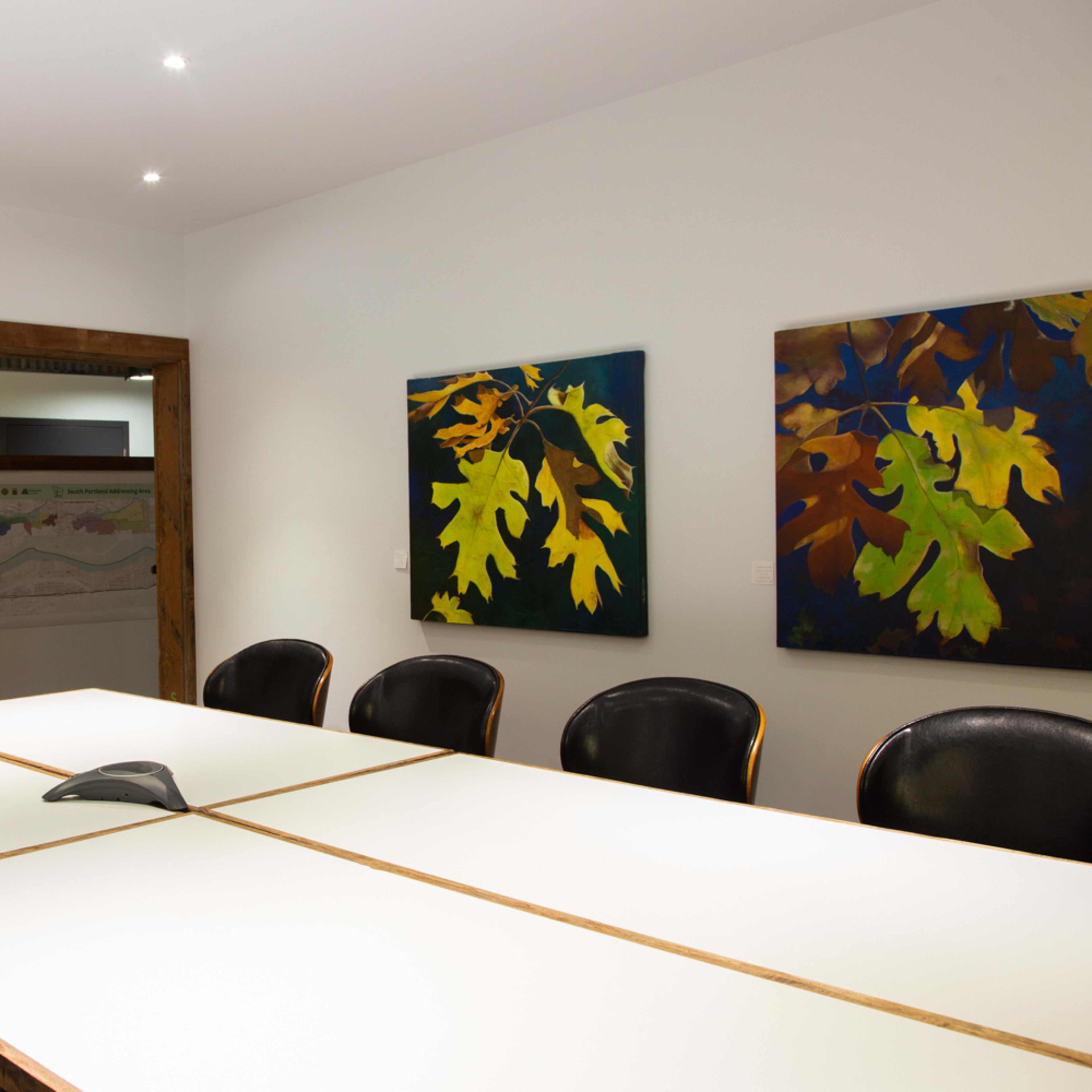 A conference room features a long white table surrounded by black chairs, with a wall-mounted TV and two large colorful paintings depicting leaves.