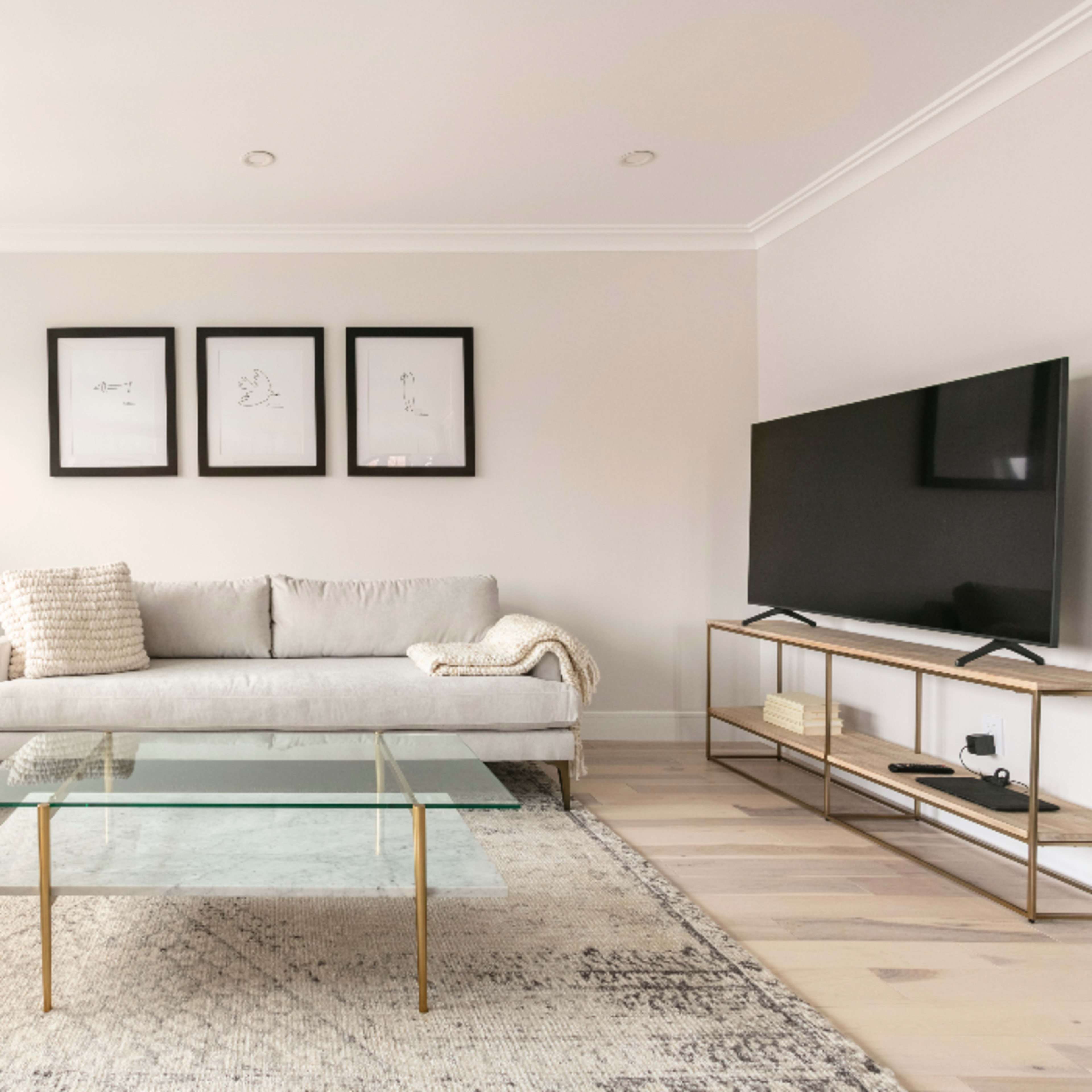 A modern living room features a light-colored sofa, a glass coffee table, a television on a stand, and framed artwork on the walls.