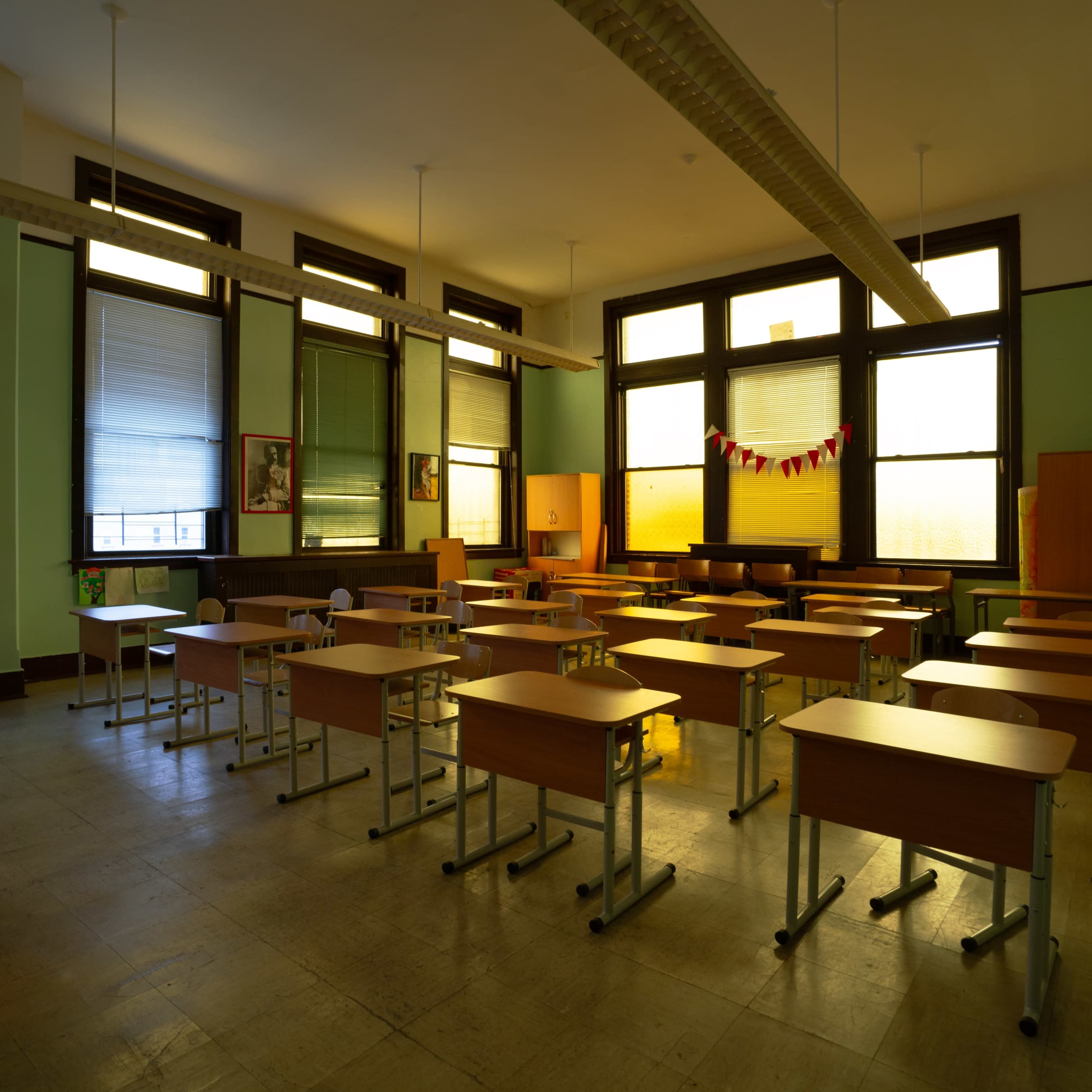 A vacant classroom features rows of wooden desks and large windows, with green walls and minimal decorations.