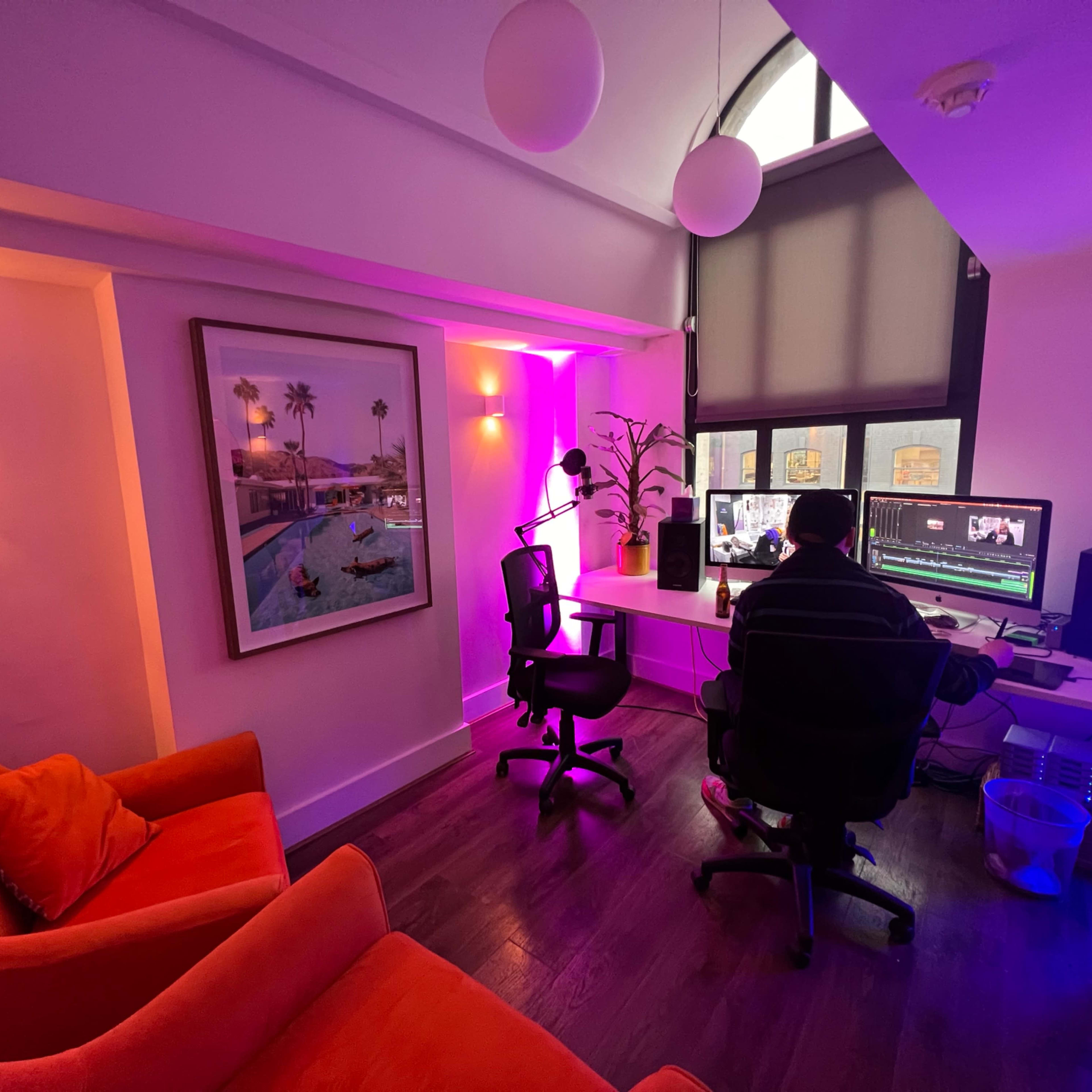 A brightly lit workspace with a person editing video on a computer, surrounded by orange seating and colorful ambient lighting.
