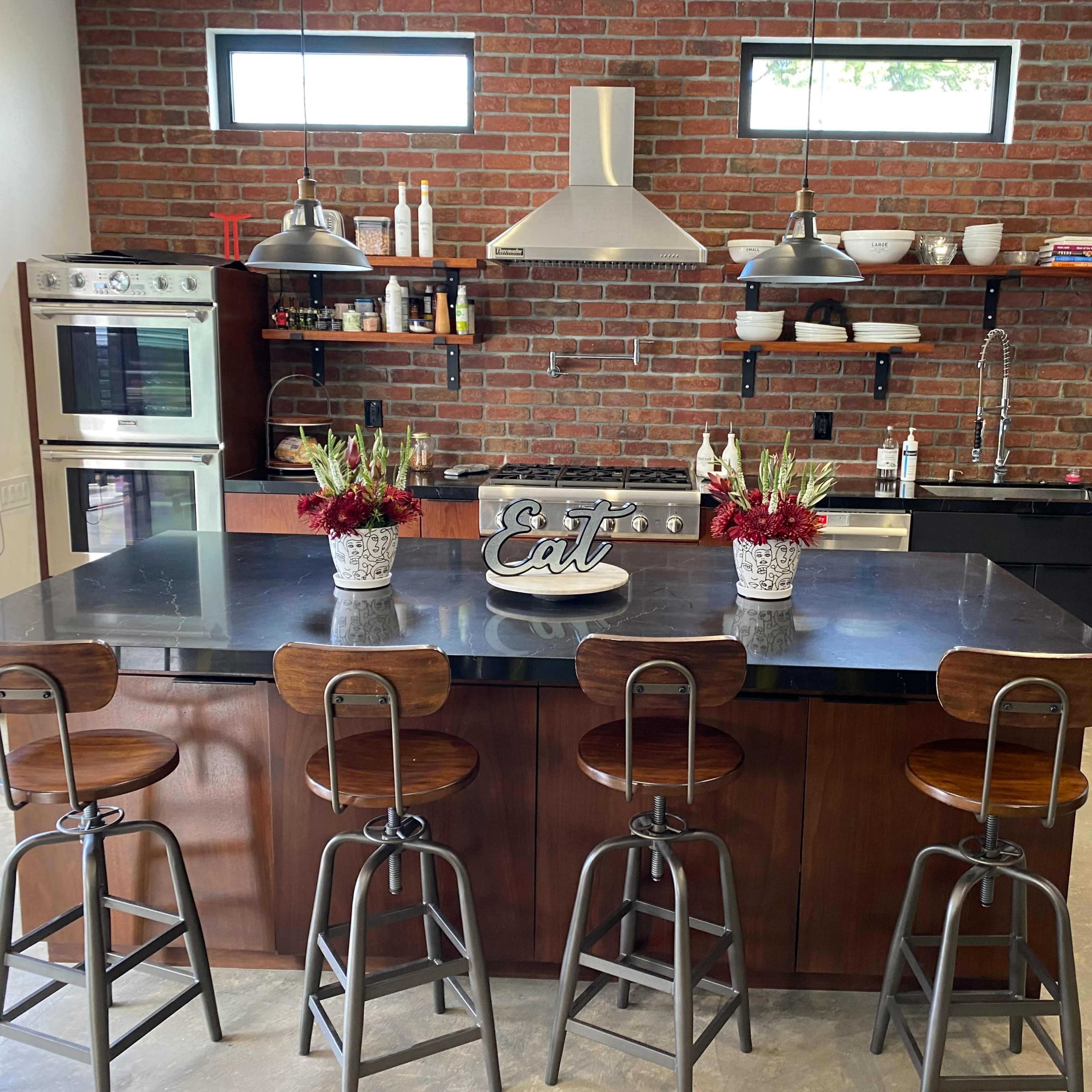 A modern kitchen features a large island with four metal stools, brick walls, and open shelving displaying various kitchen items.