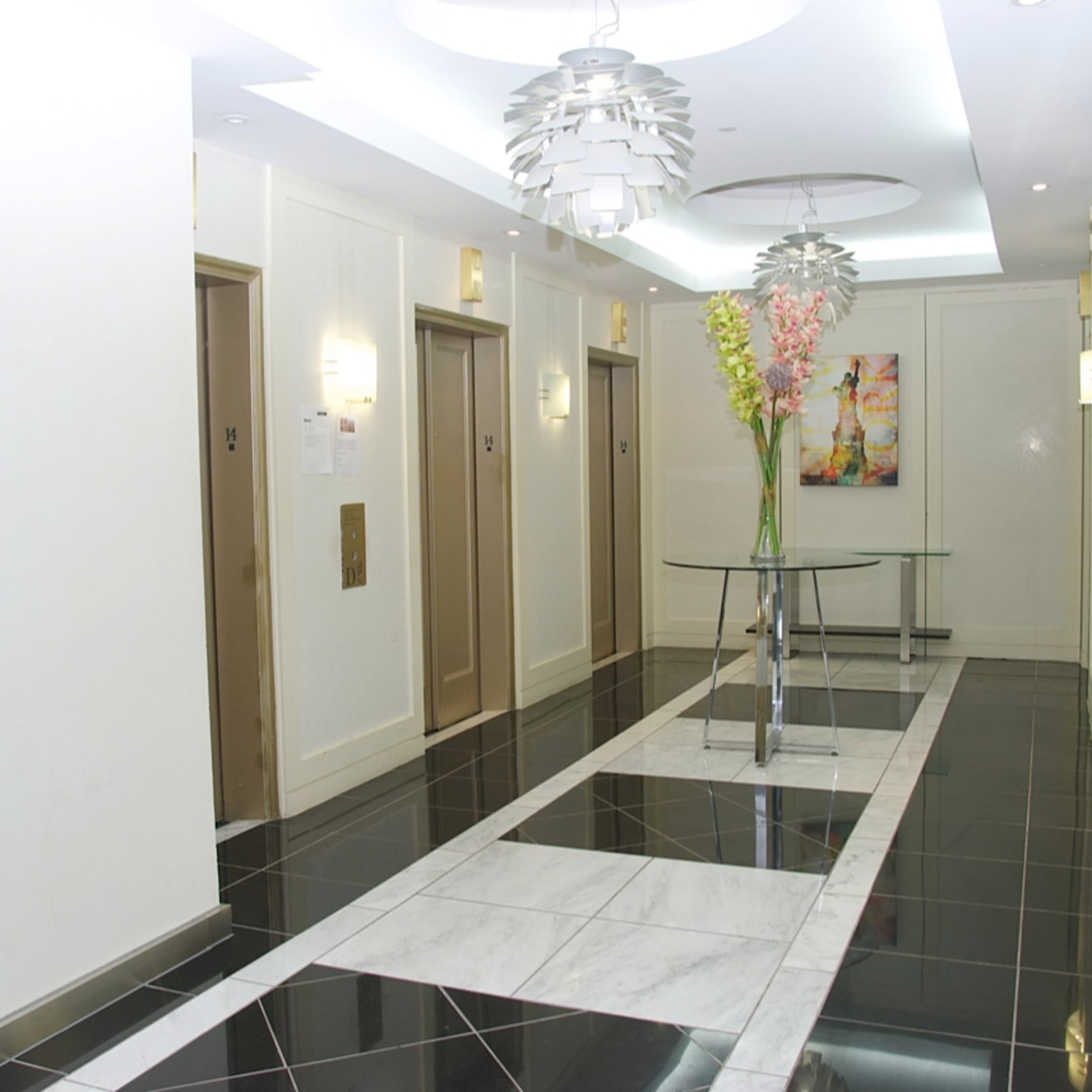 A modern hotel corridor with elevator doors on either side, a glass table with flowers in the center, and a black and white tile floor.
