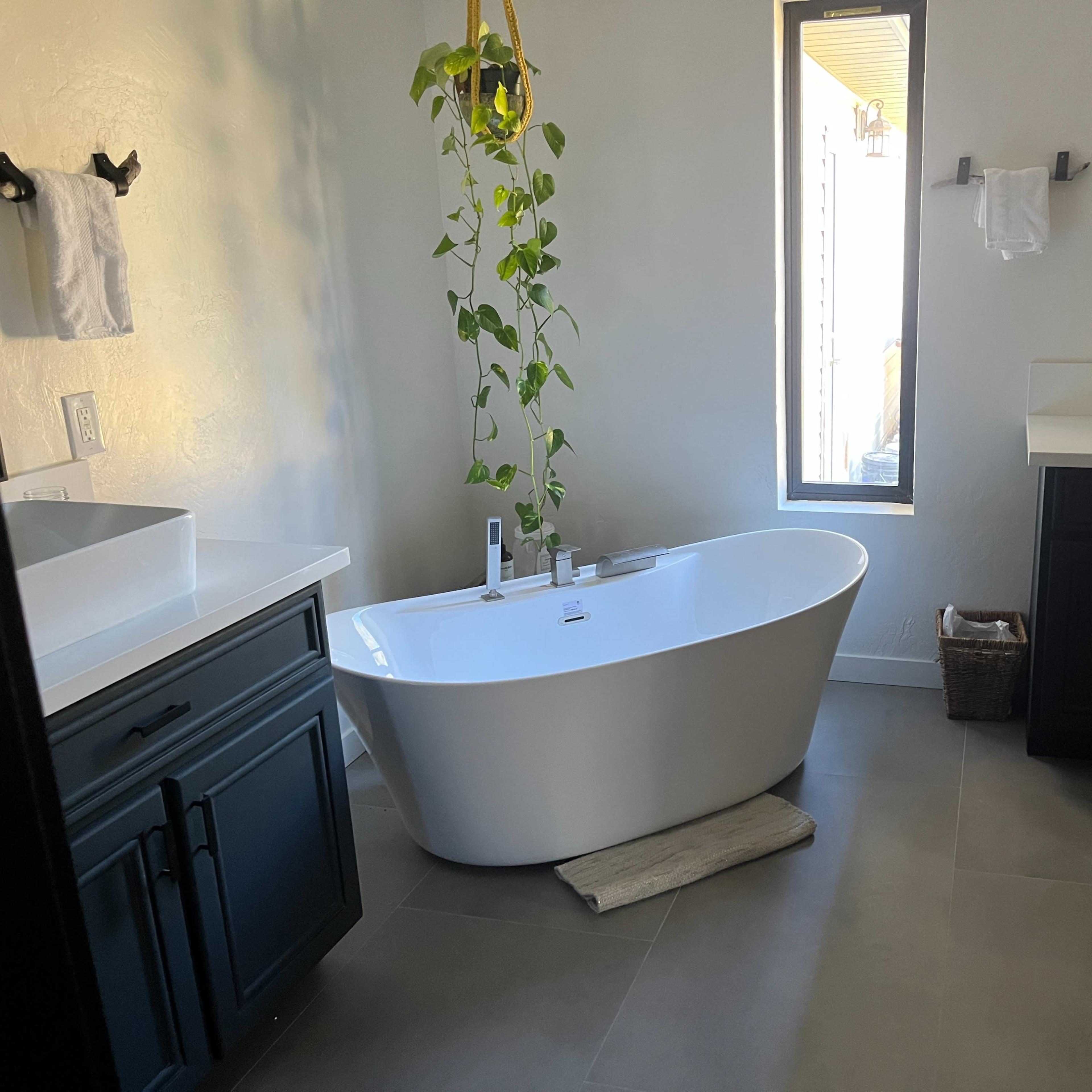 A modern bathroom features a freestanding bathtub, a large mirror, and a sink with dark cabinetry, illuminated by natural light from a window.