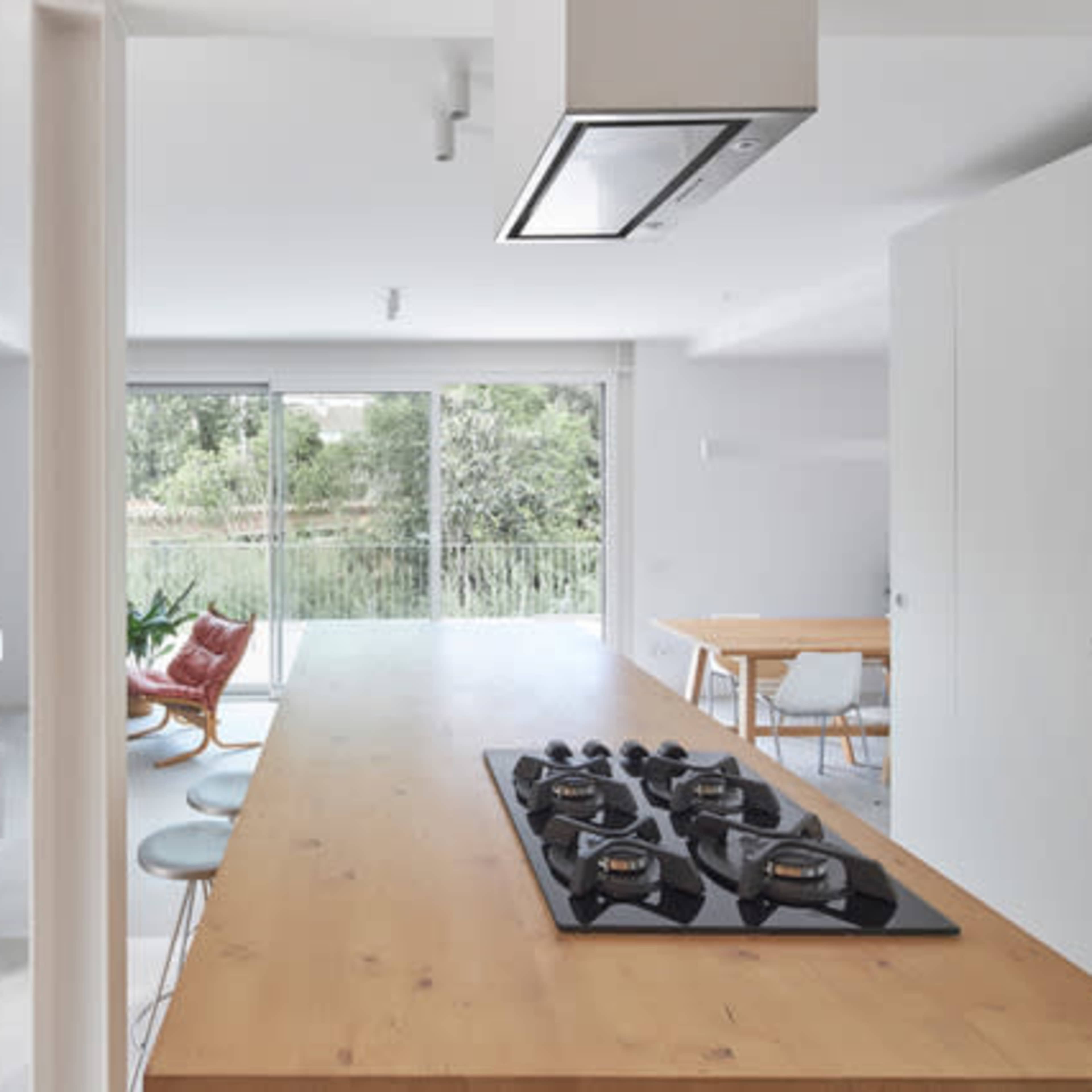 A modern kitchen with a wooden island featuring a gas stove, surrounded by white cabinetry and an open layout leading to a dining area with large windows overlooking greenery.