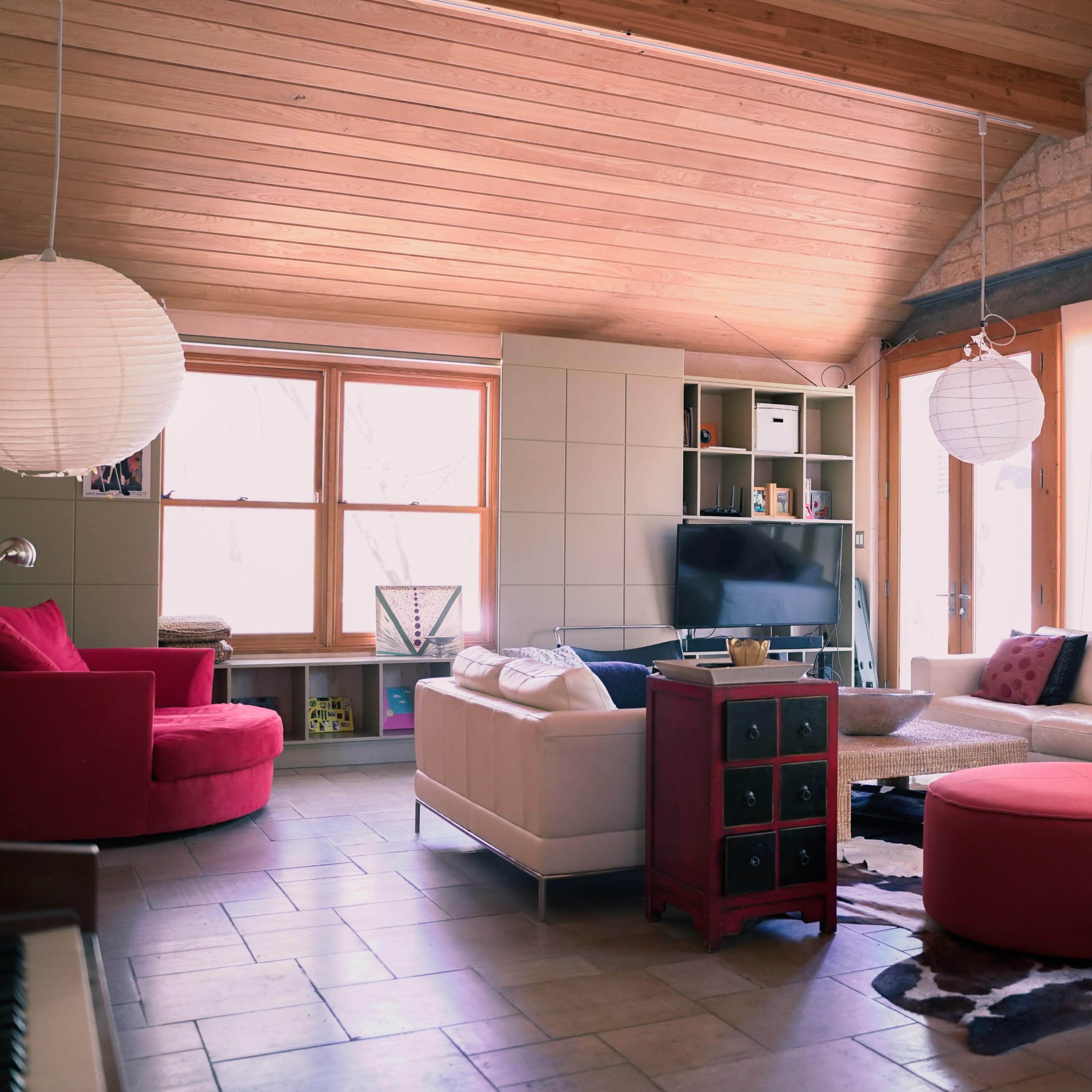 A modern living room features a mix of seating options, including a red chair, light-colored sofas, and a round ottoman, with large windows allowing natural light to filter in.