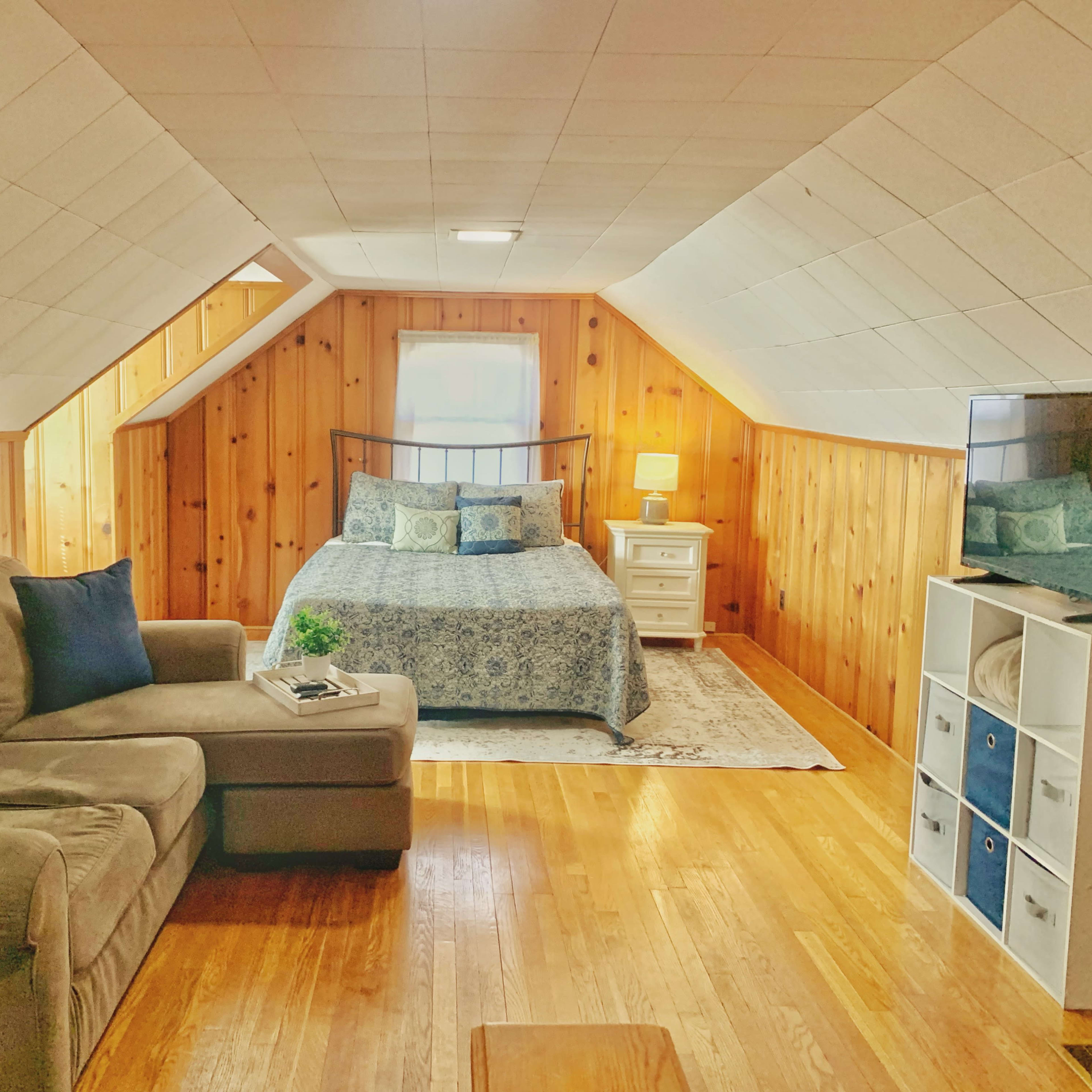 The room features a cozy bedroom with a floral-patterned bedspread, a small nightstand with a lamp, a sofa, and a television against wooden paneled walls.