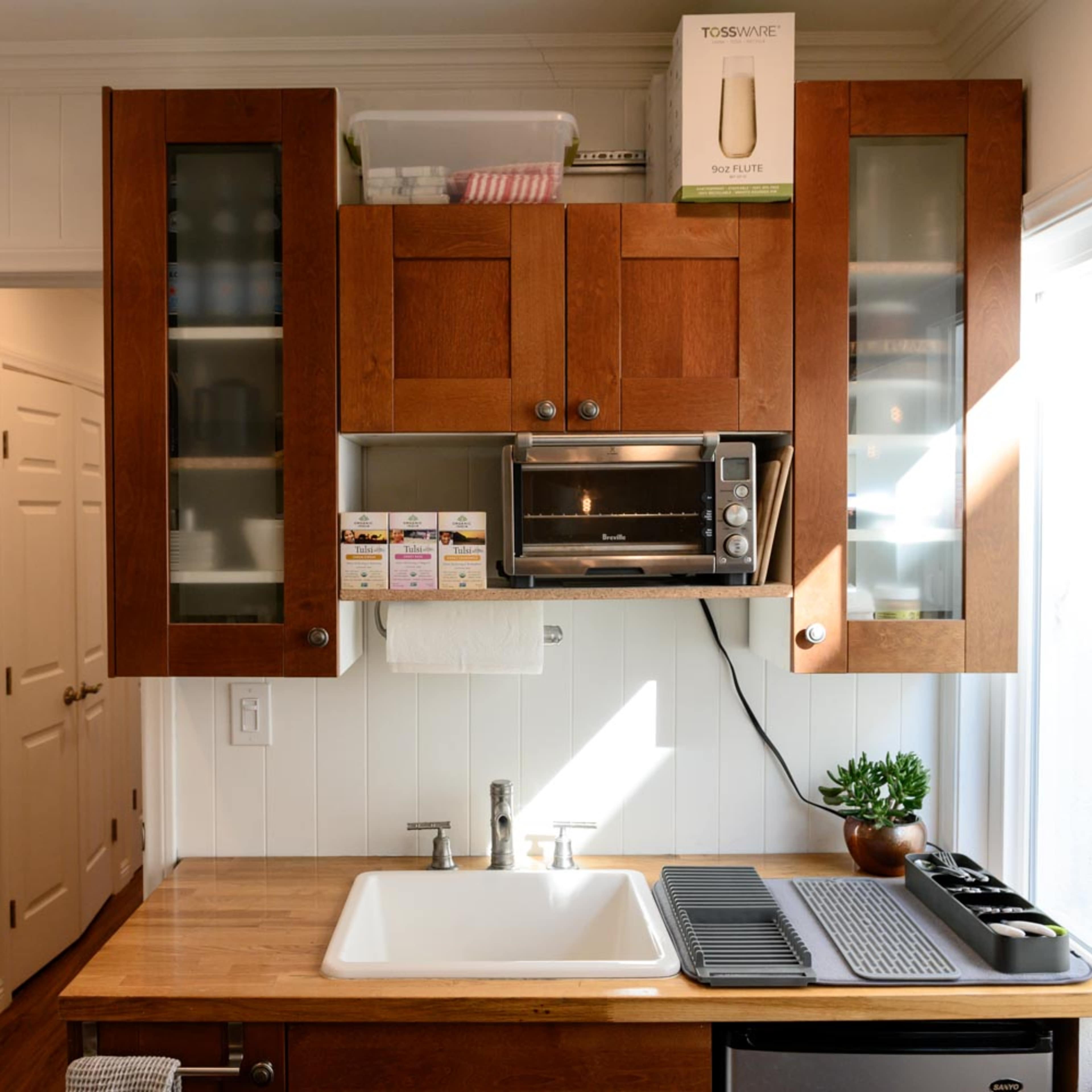 A modern kitchen with wooden cabinets, a stainless steel microwave above a farmhouse sink, and a countertop with a dish drying rack and small plant.