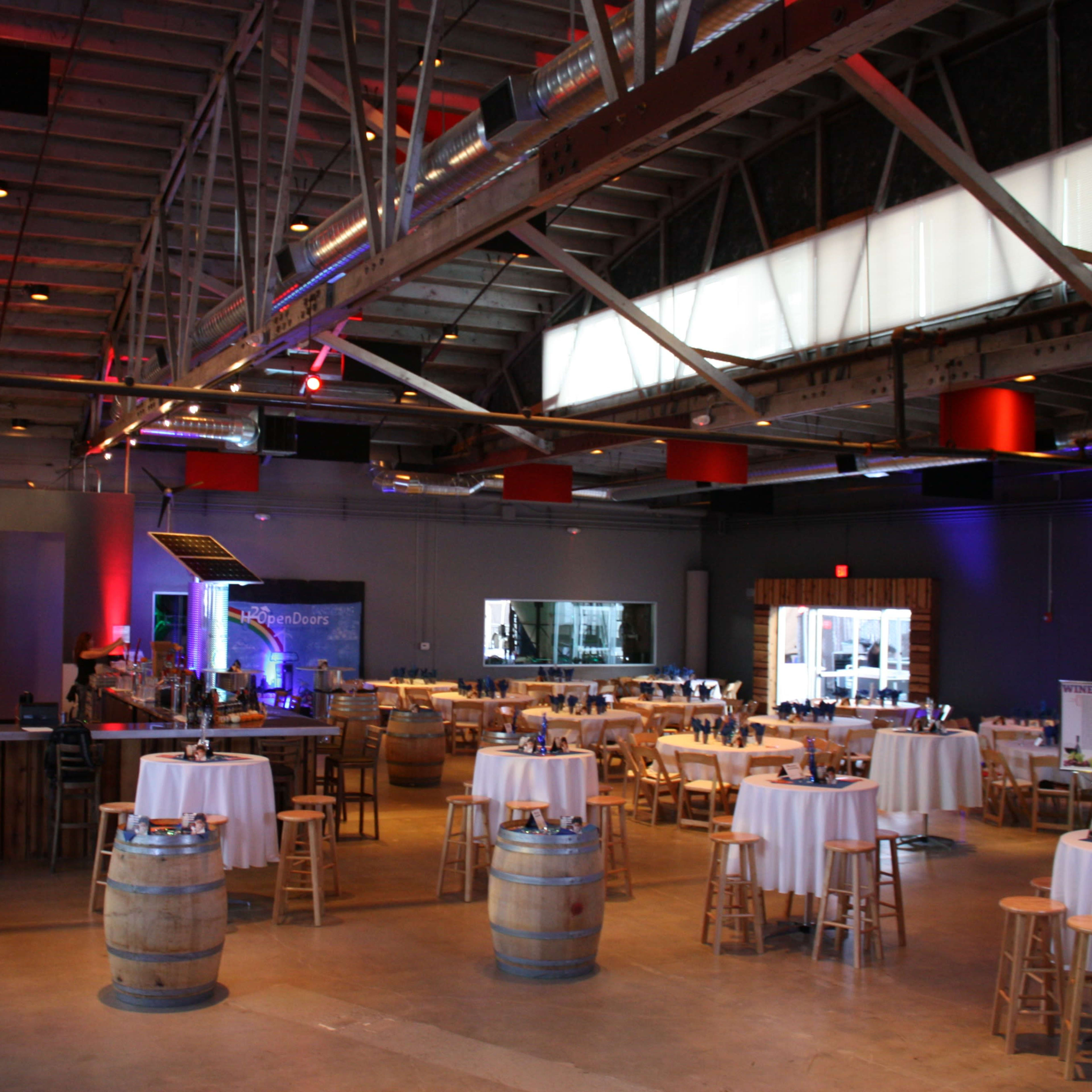 A spacious event venue with circular tables set for dining, surrounded by bar stools, and a bar area in the back.