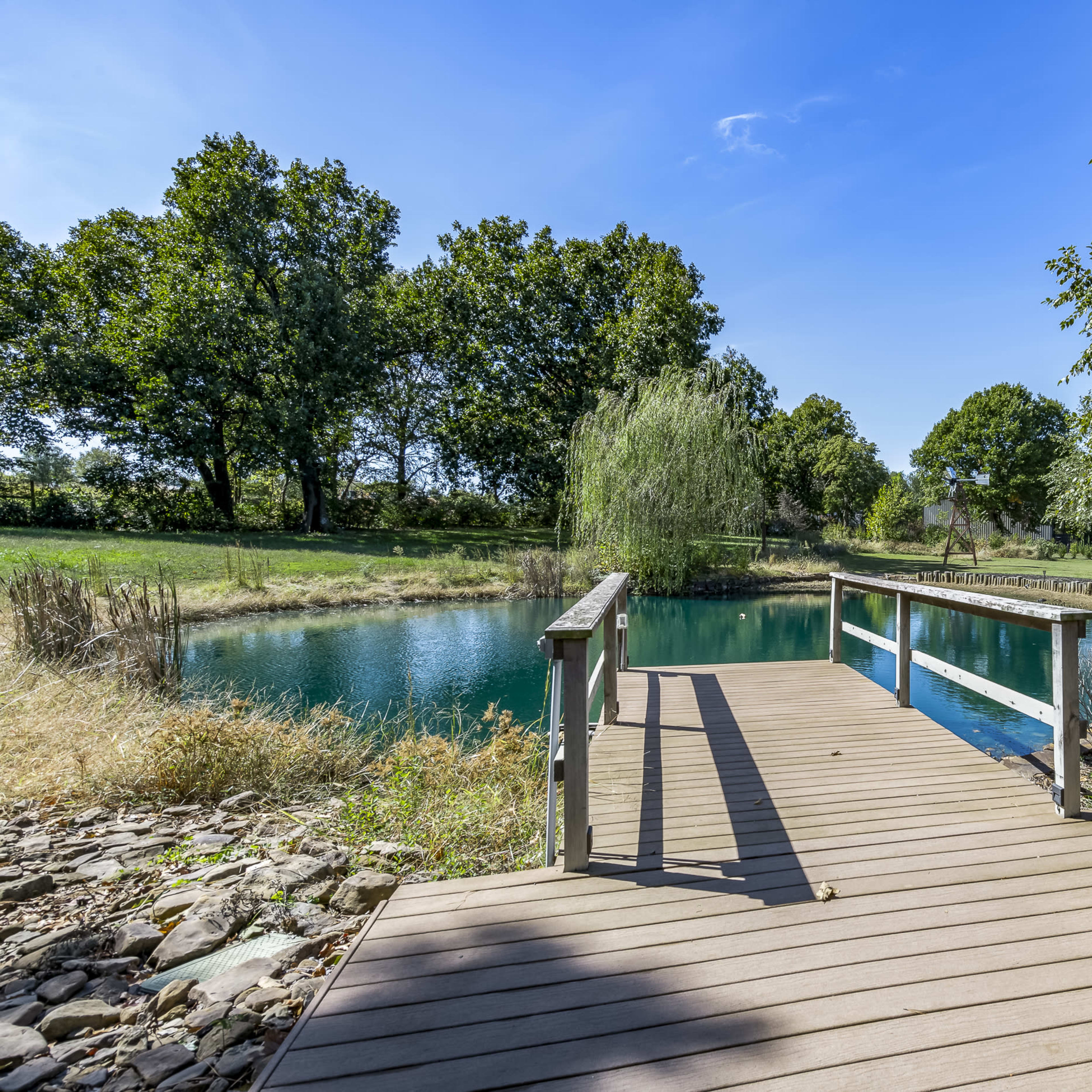 A wooden dock extends over a calm pond surrounded by trees and grassy areas under a clear blue sky.