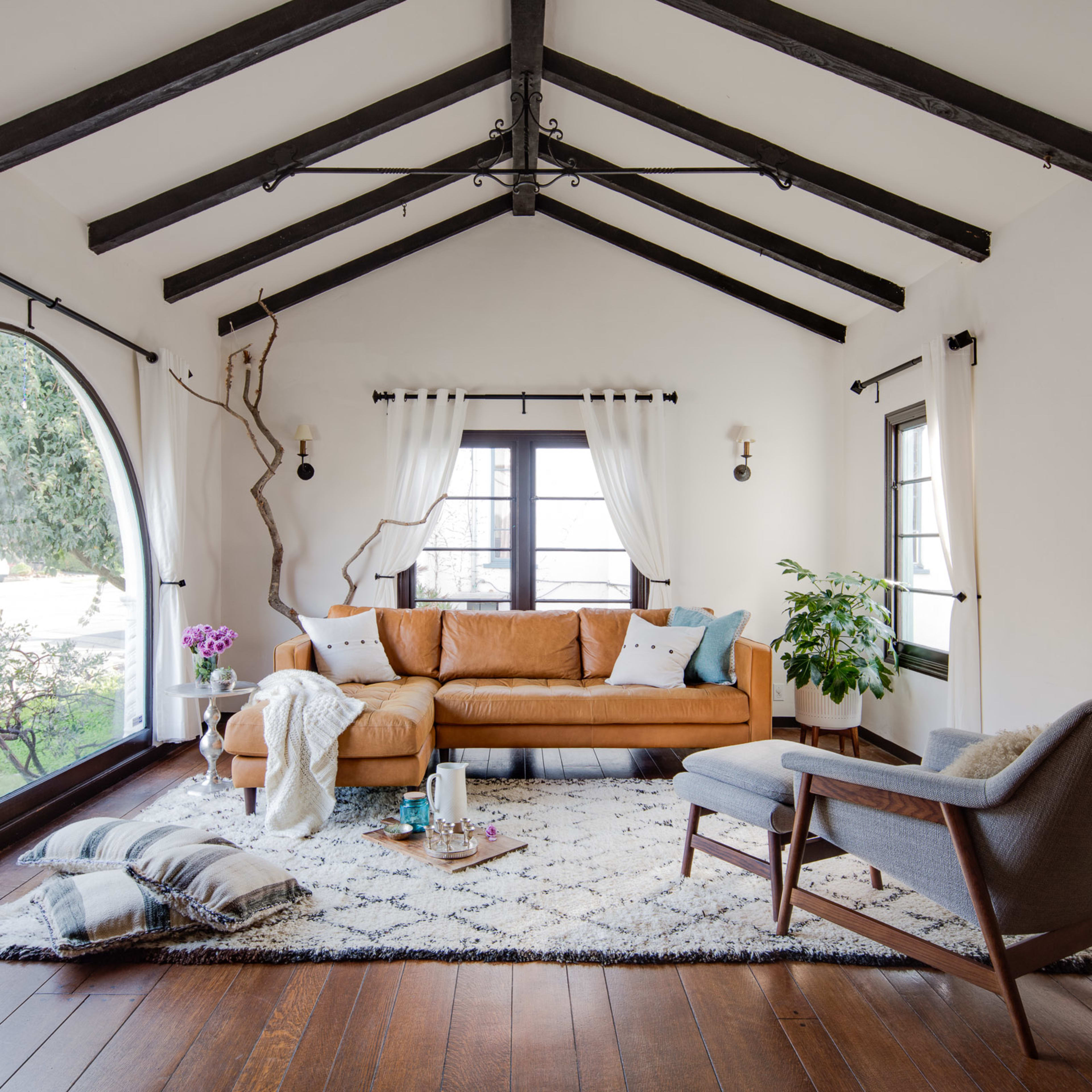 A living room with a brown leather sofa, a gray armchair, and a large area rug, illuminated by natural light through two windows.