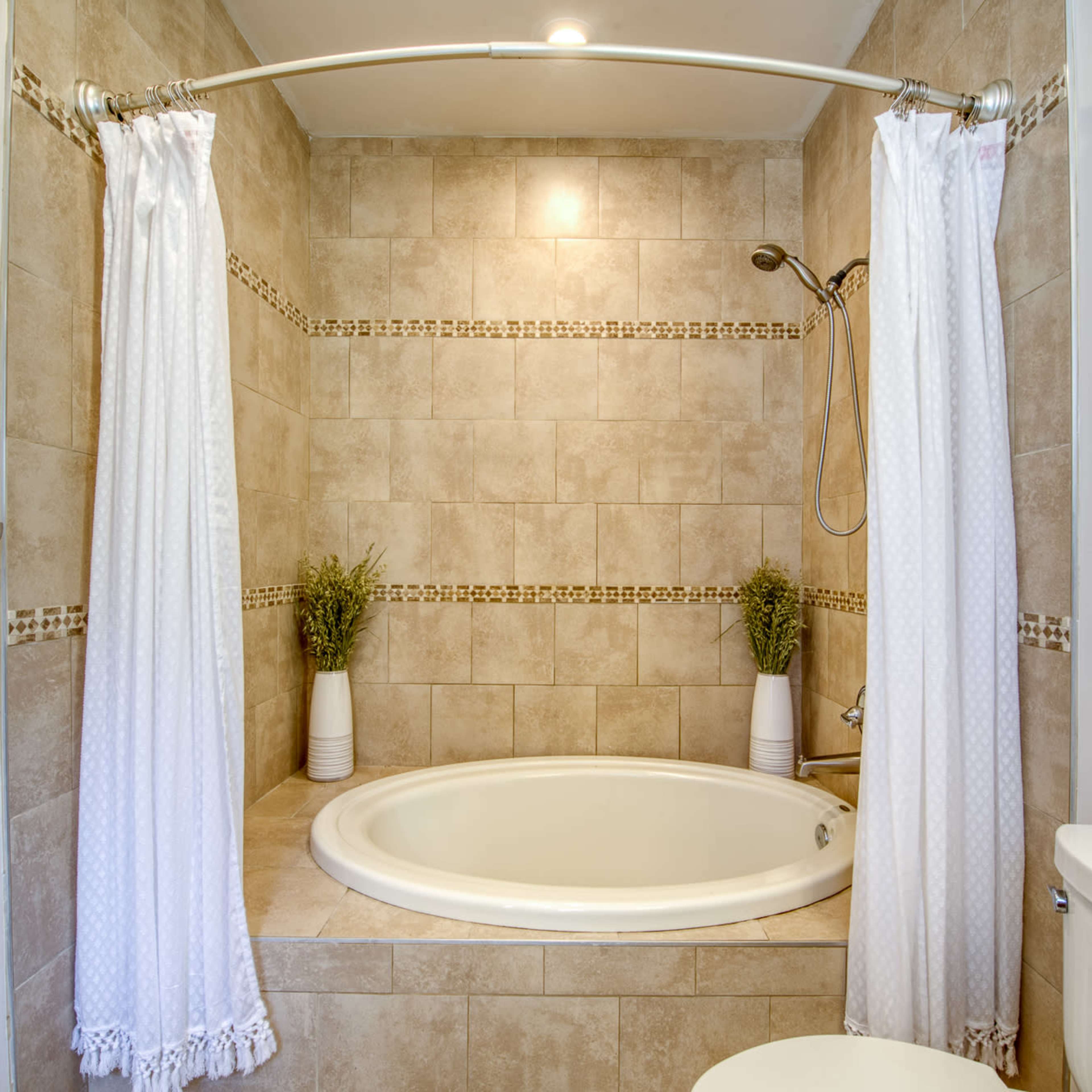 A small bathroom features a round soaking tub surrounded by tiled walls, with a showerhead and white curtains on either side.