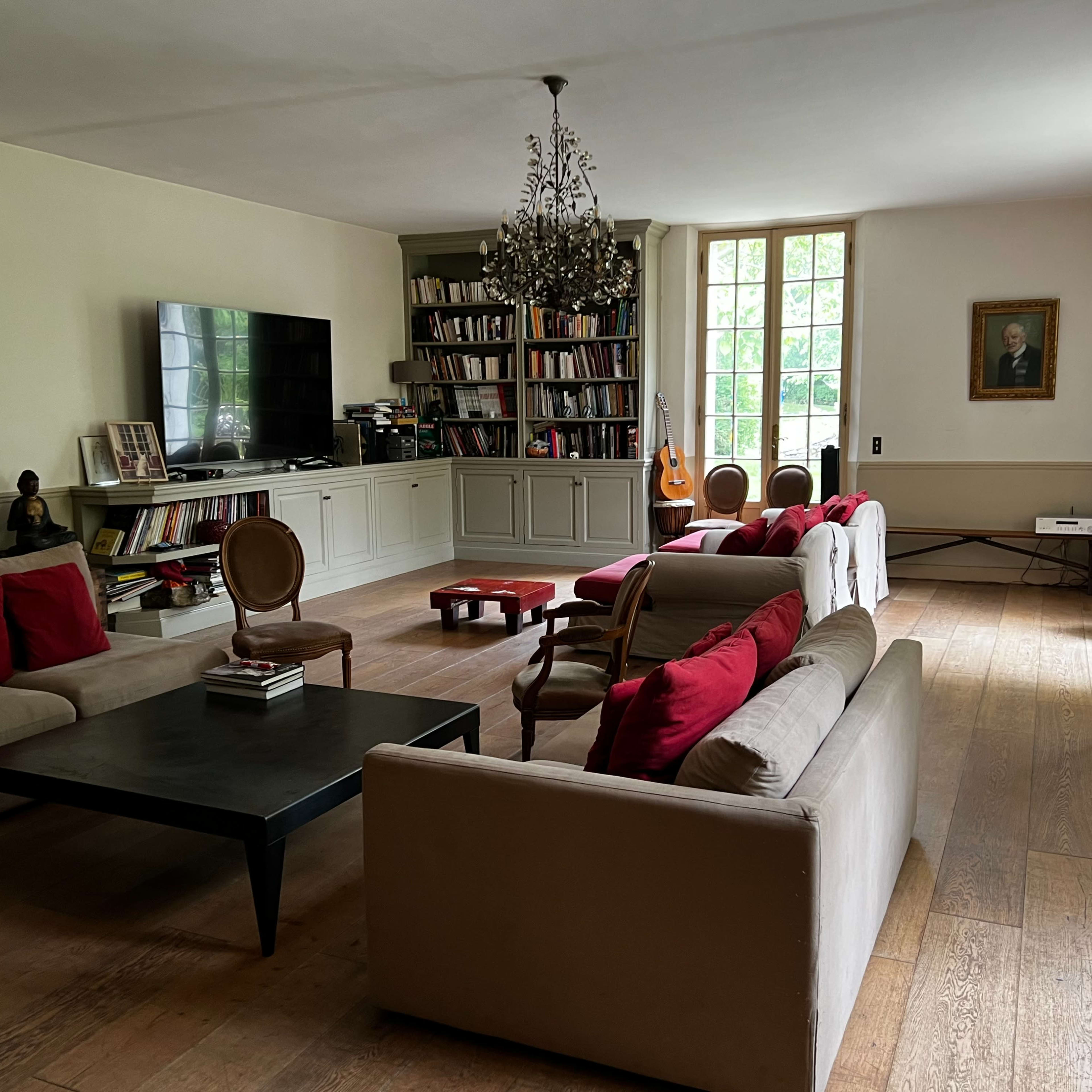 The room features a spacious living area with multiple seating arrangements, a television, bookshelves, and a large window allowing natural light.