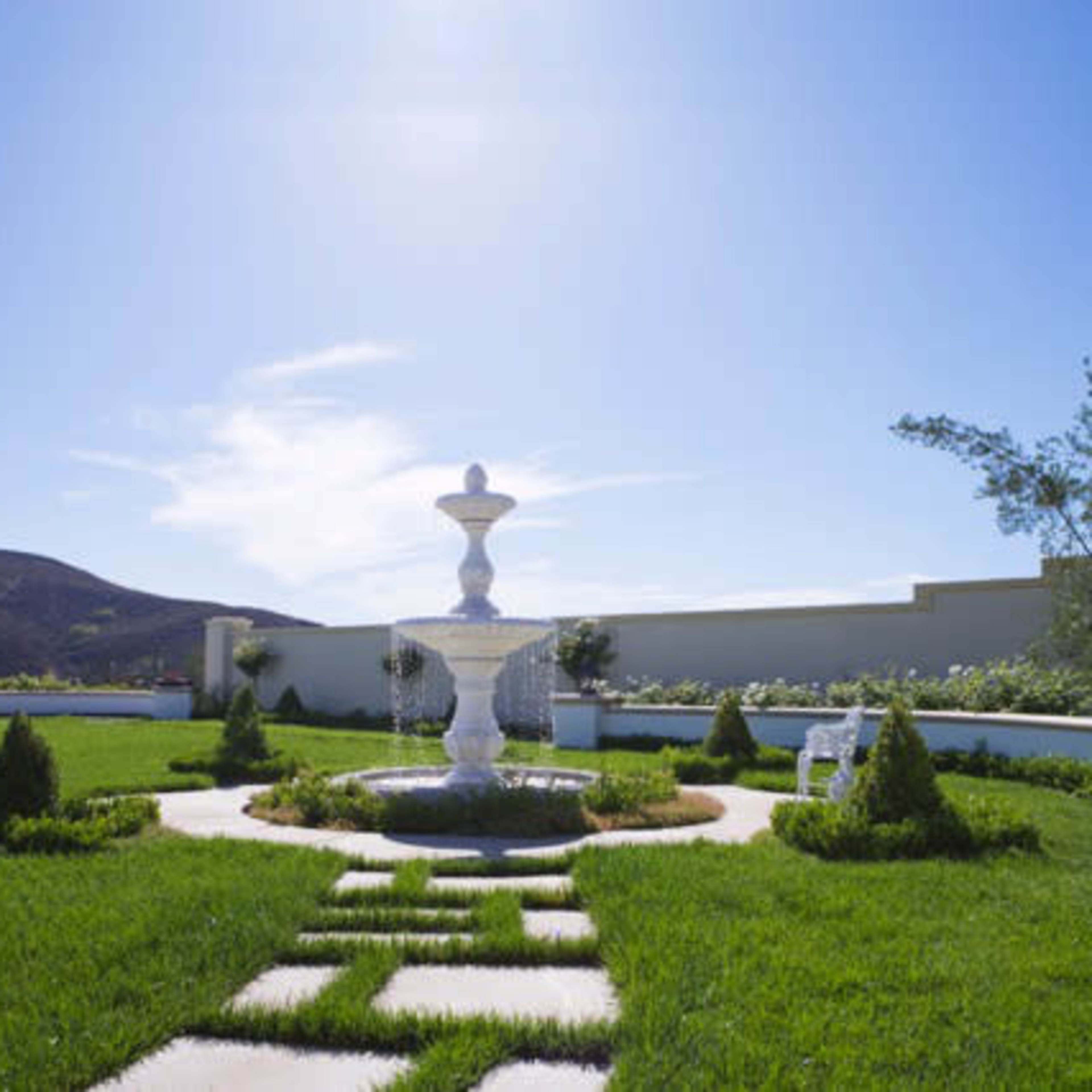 A white fountain sits at the center of a manicured lawn surrounded by neatly trimmed hedges and a view of a hillside under a clear blue sky.