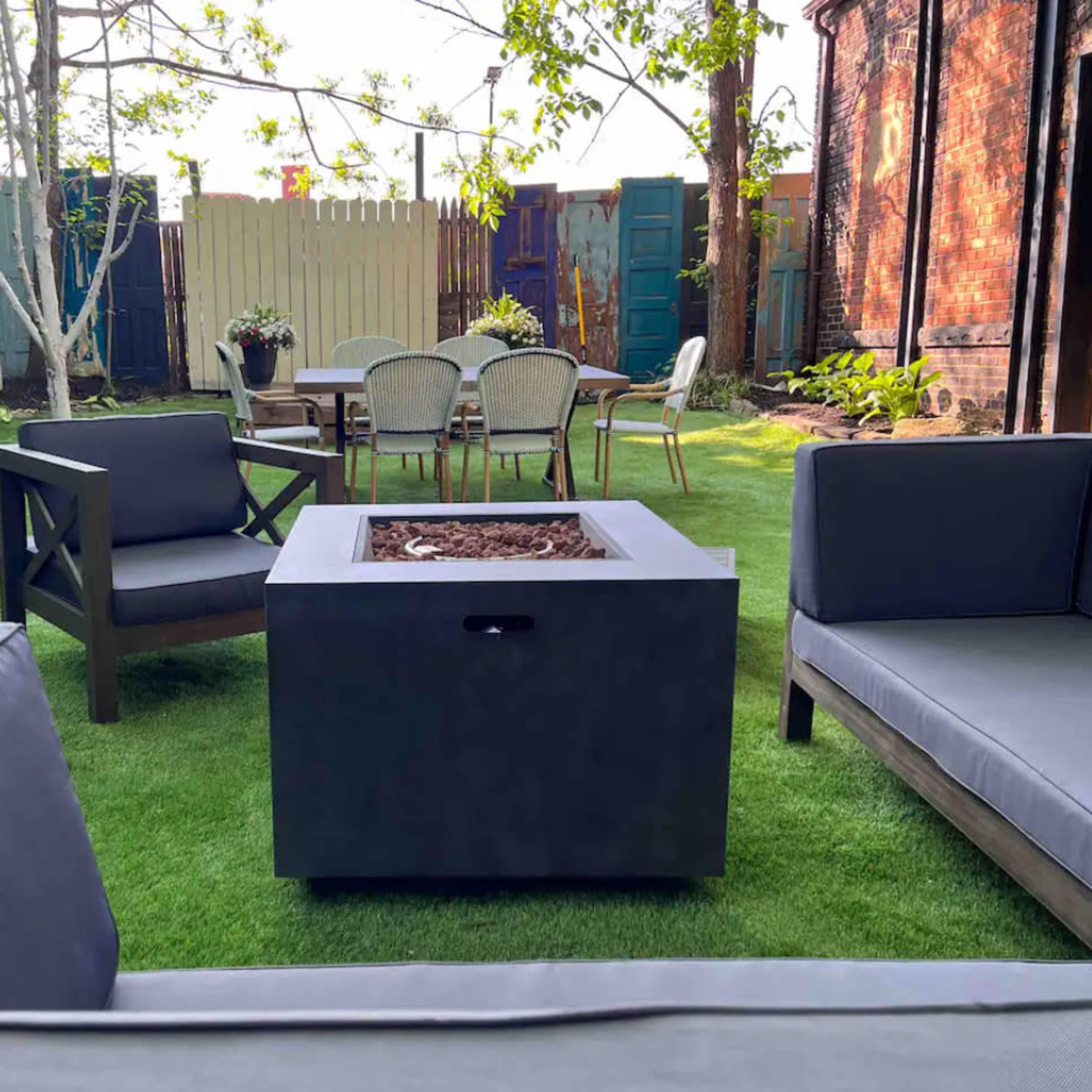 A patio area features several seating arrangements around a central fire pit, with greenery and outdoor furniture in the background.