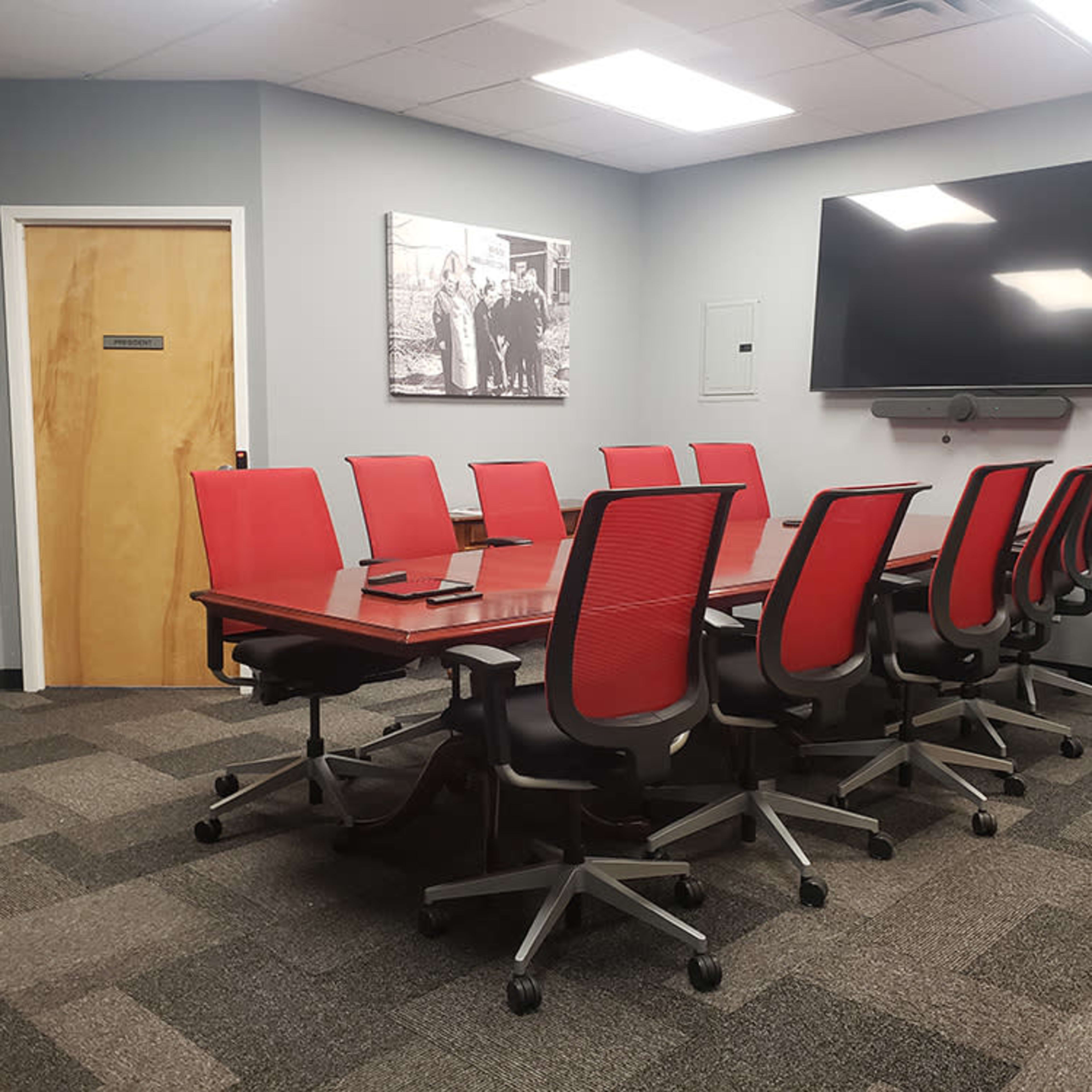 A modern conference room with a large table surrounded by red chairs, a wall-mounted TV, and a door leading to another area.