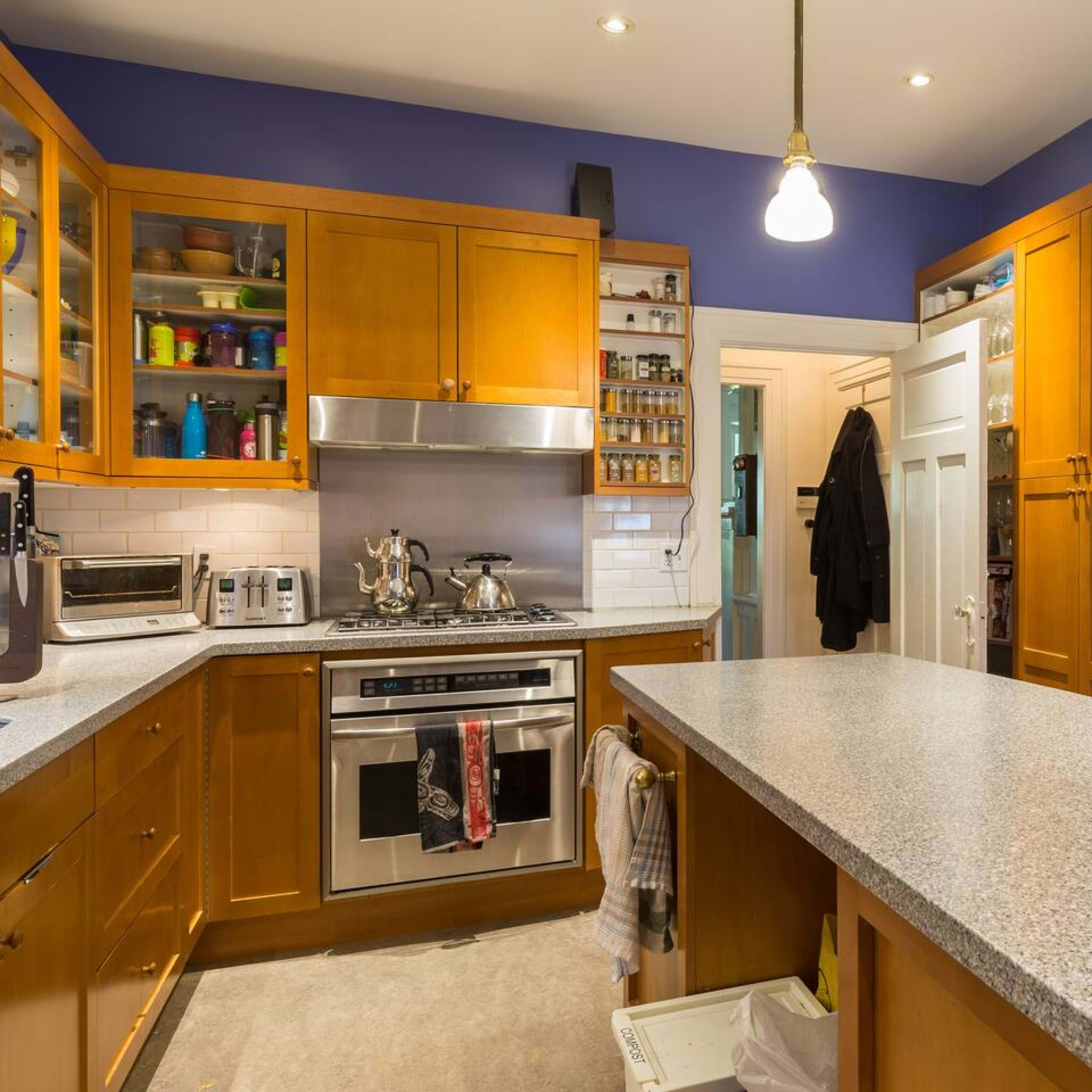 A compact kitchen with wooden cabinets, a stainless steel oven, and a central countertop illuminated by a hanging light fixture.