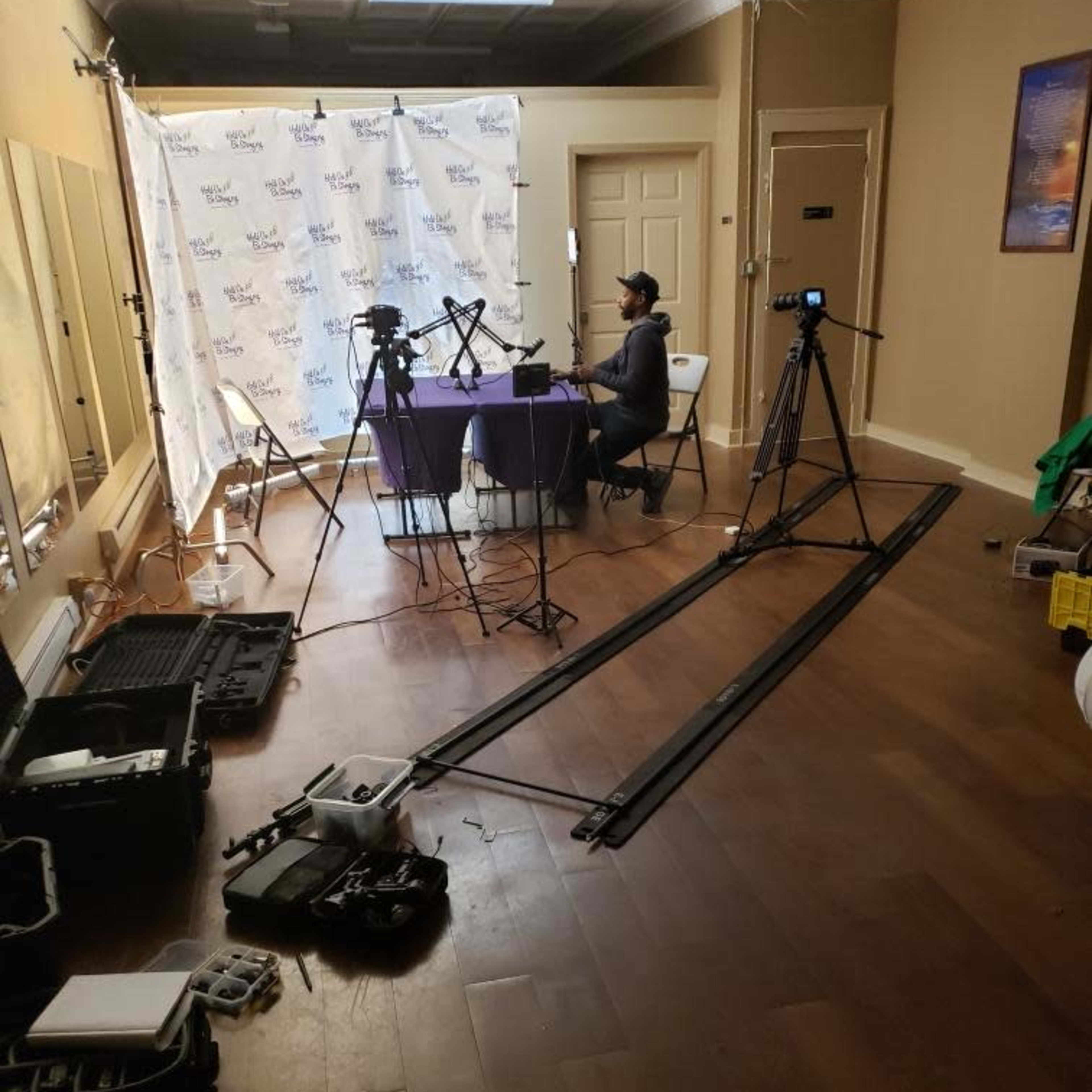 A person sits at a table in a room set up for video production, surrounded by cameras, equipment, and a backdrop with a logo.