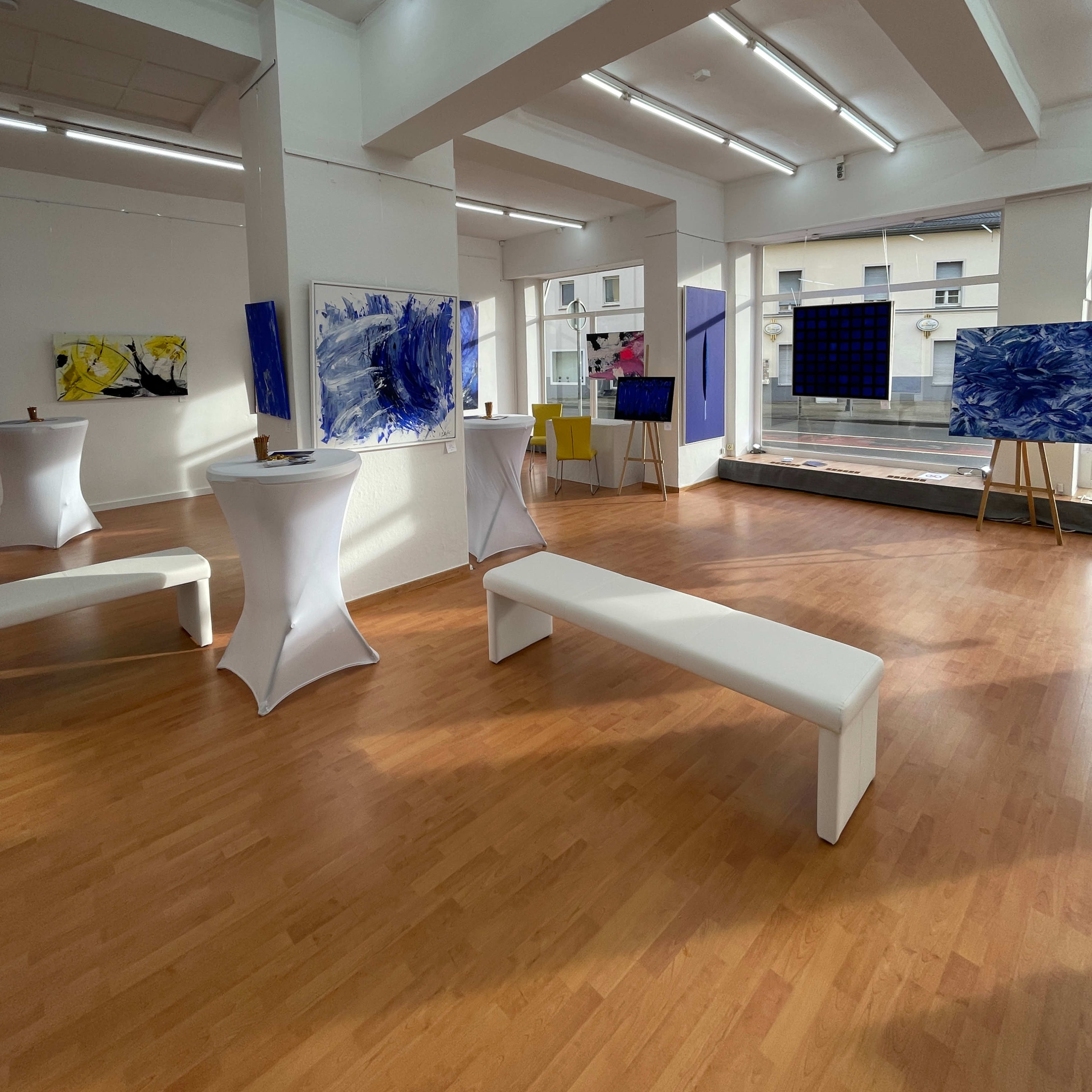 An art gallery featuring abstract paintings on the walls, with white benches and table setups arranged in the space.