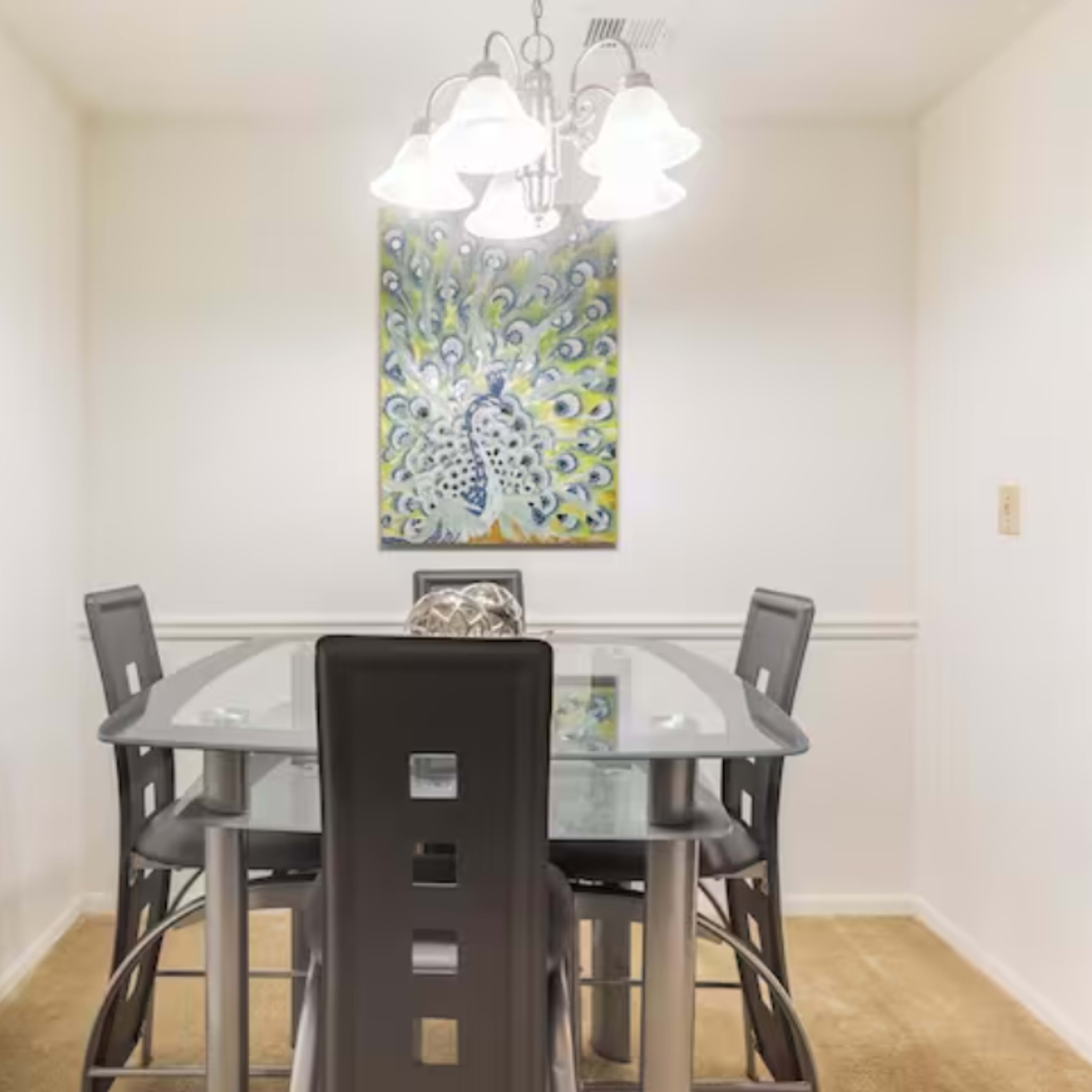 A glass dining table with four black chairs is illuminated by a pendant light, while a colorful artwork hangs on the wall.