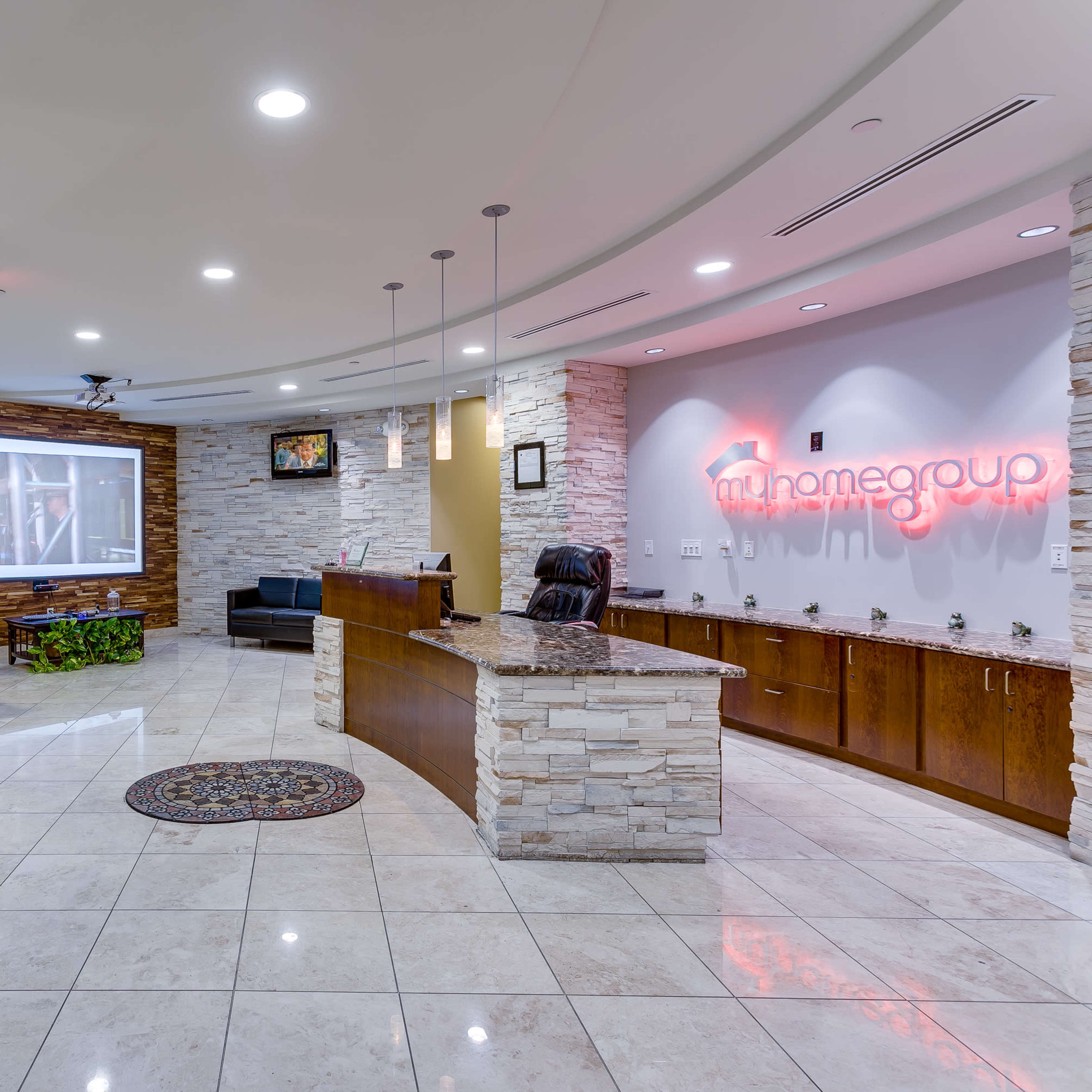 A modern reception area featuring a stone-accented front desk, comfortable seating, and a decorative rug on a tiled floor, with branding displayed on the wall.