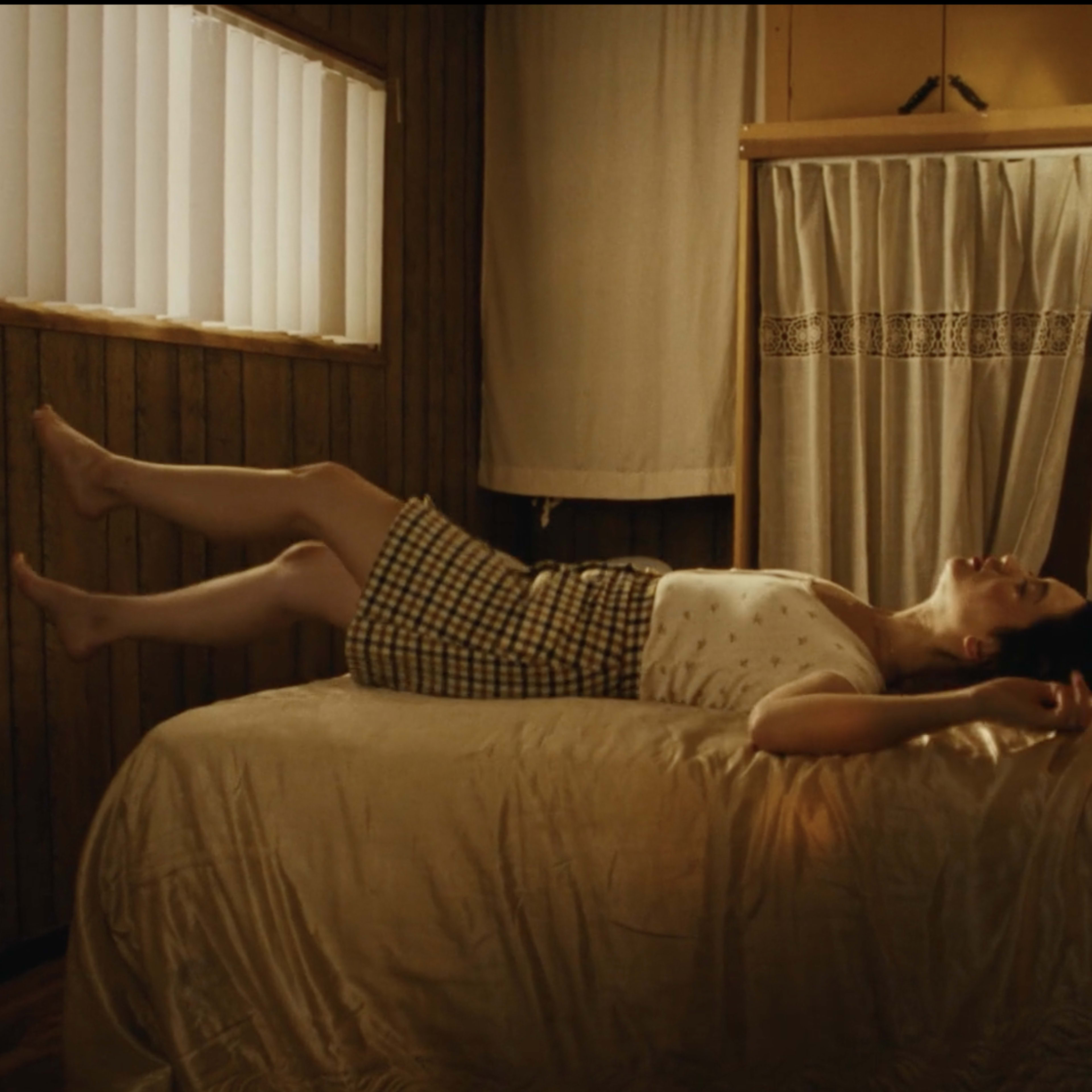 A woman lies on a bed in a wooden room, with her legs raised and eyes closed.