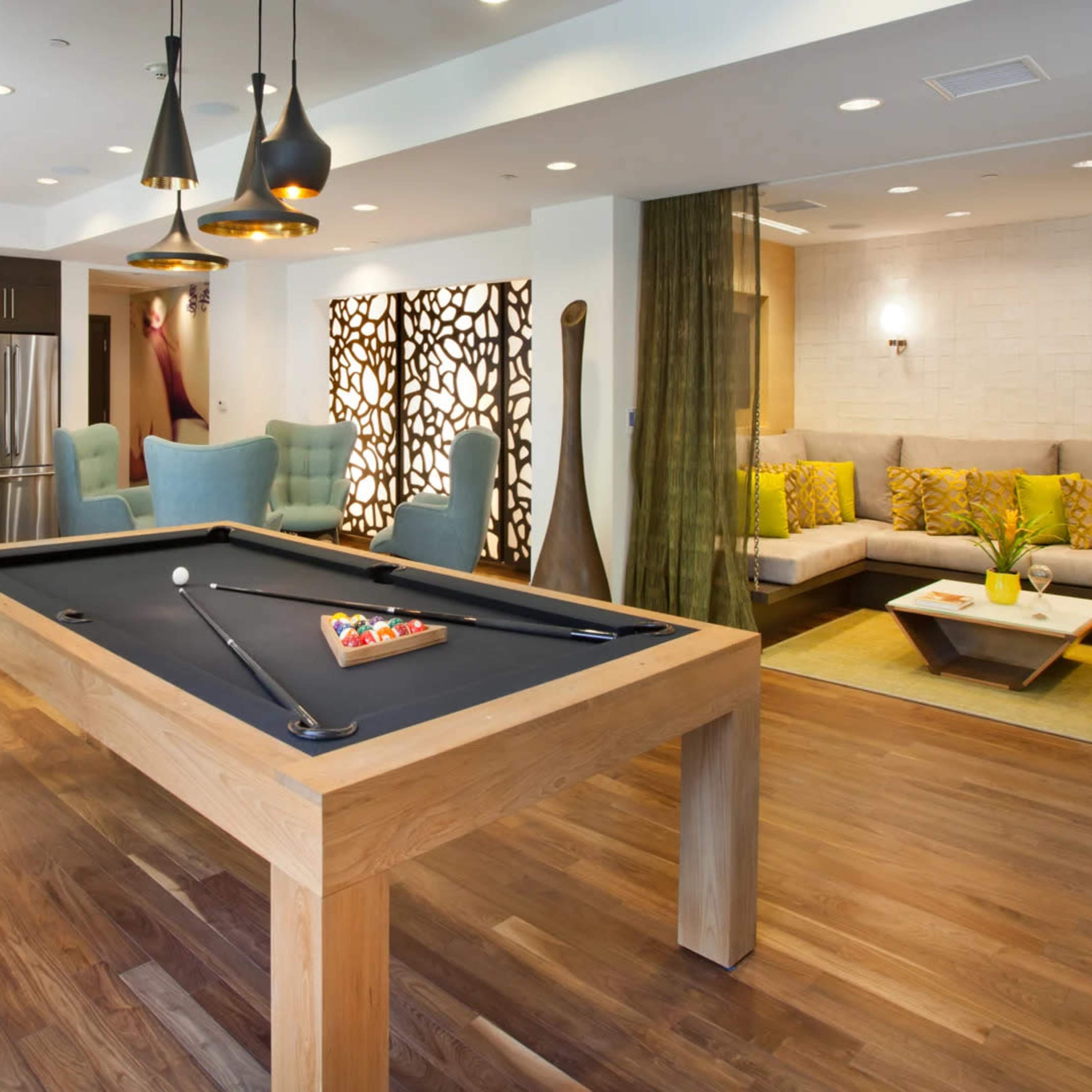 A modern recreational room features a pool table, seating area with a sofa and chairs, and a kitchenette in the background.