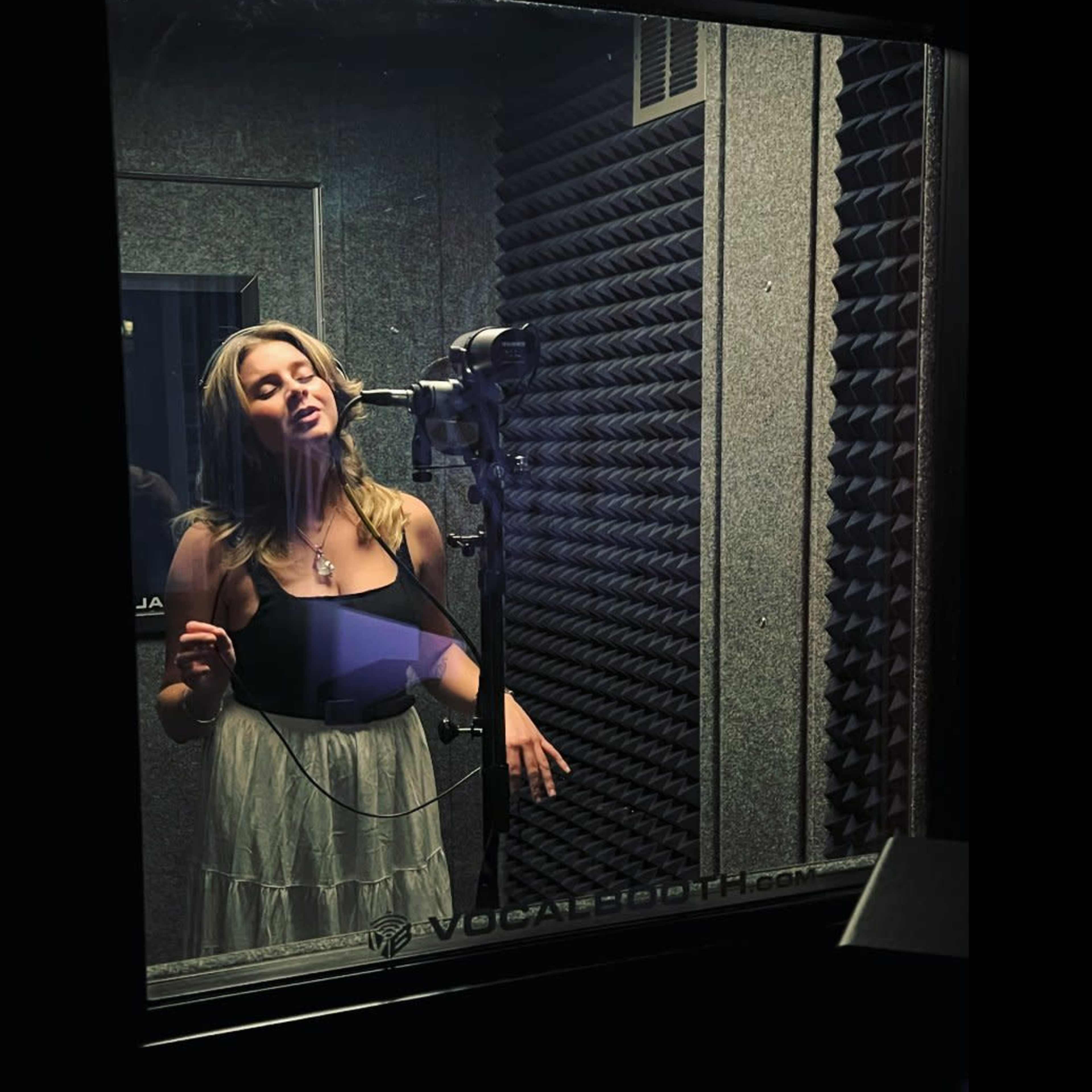 A woman stands in a soundproof recording booth, singing into a microphone.