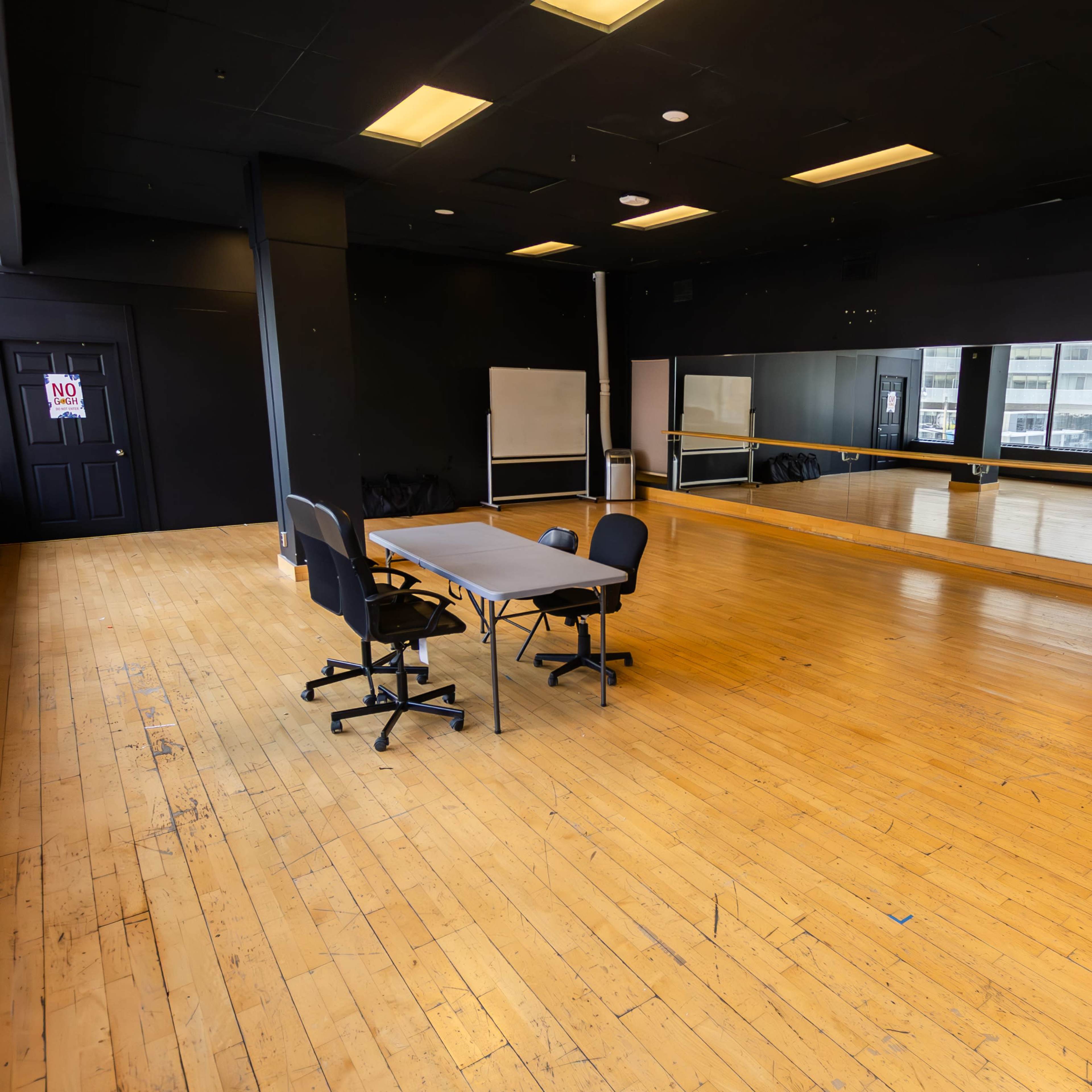 An empty room with wooden flooring, a folding table, rolling chairs, and large mirrors along one wall.