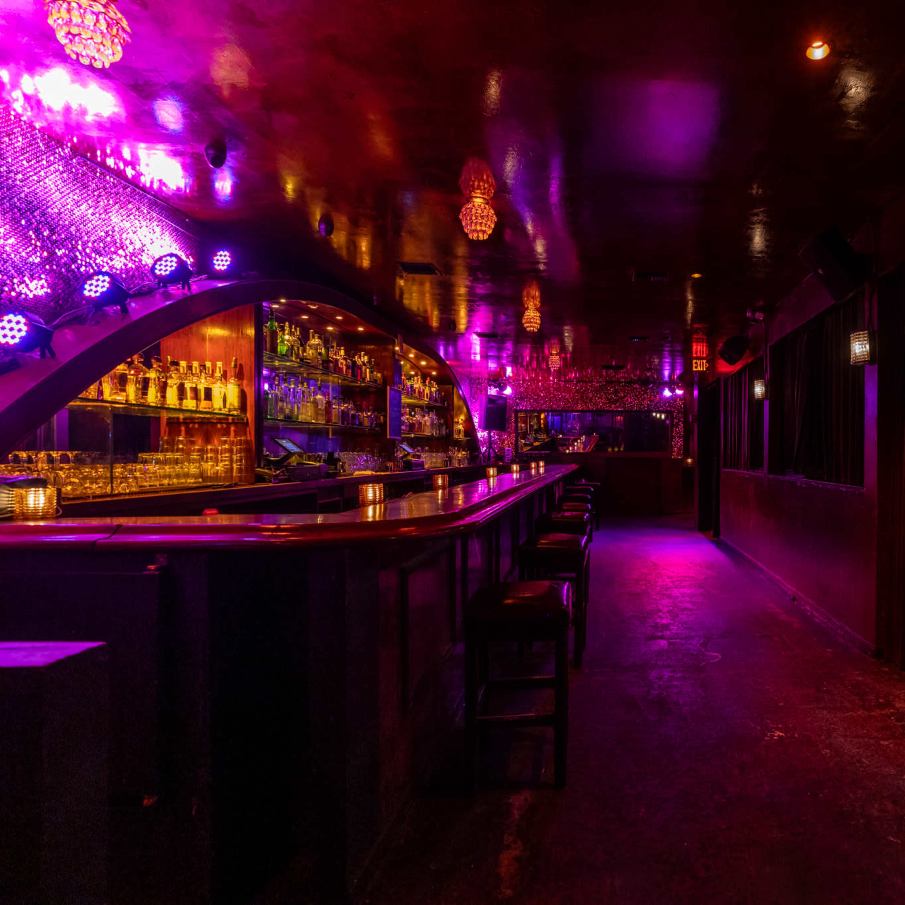 A dimly lit bar interior featuring a long bar counter, illuminated shelves of alcohol, and colorful light fixtures reflecting off the walls.