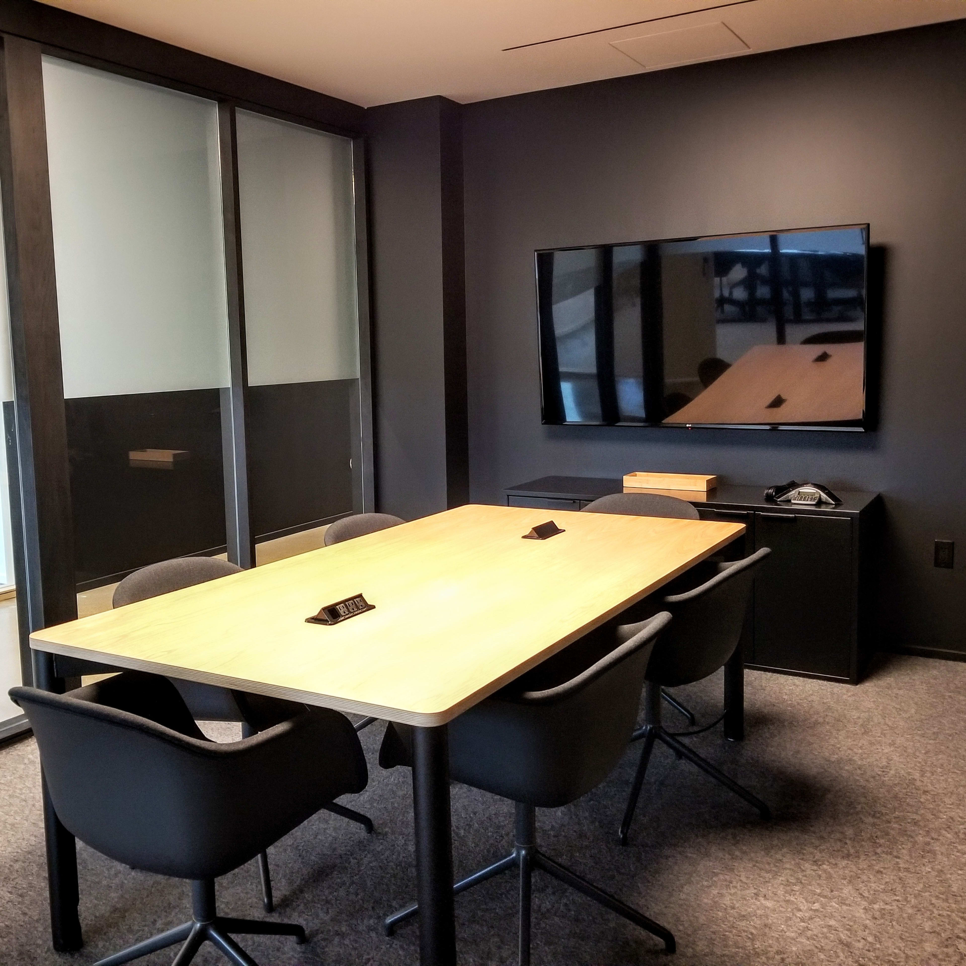 A modern conference room featuring a wooden table surrounded by four chairs, a large television on the wall, and glass partitions leading to another area.