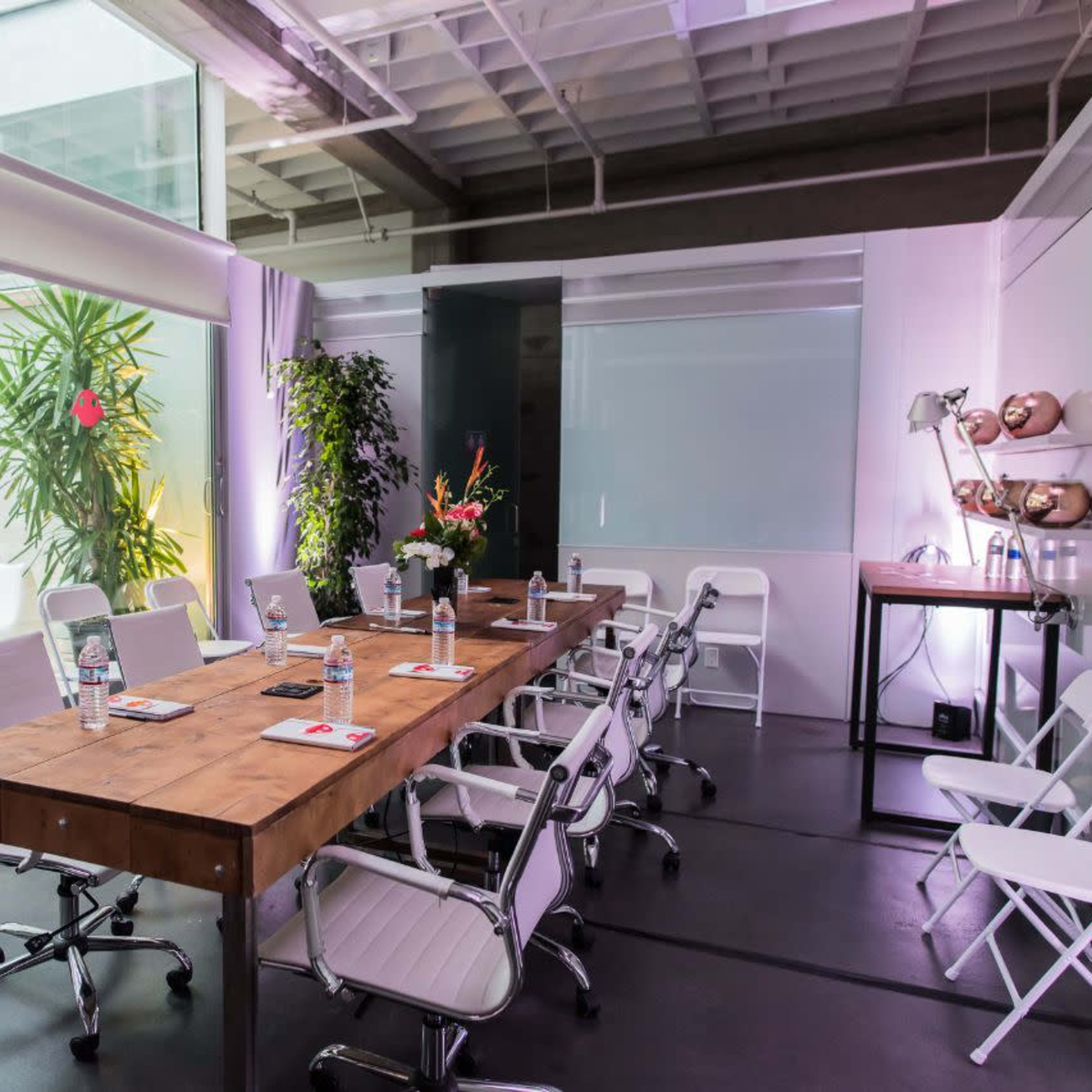 A modern conference room with a long wooden table, white chairs, and large windows overlooking greenery.
