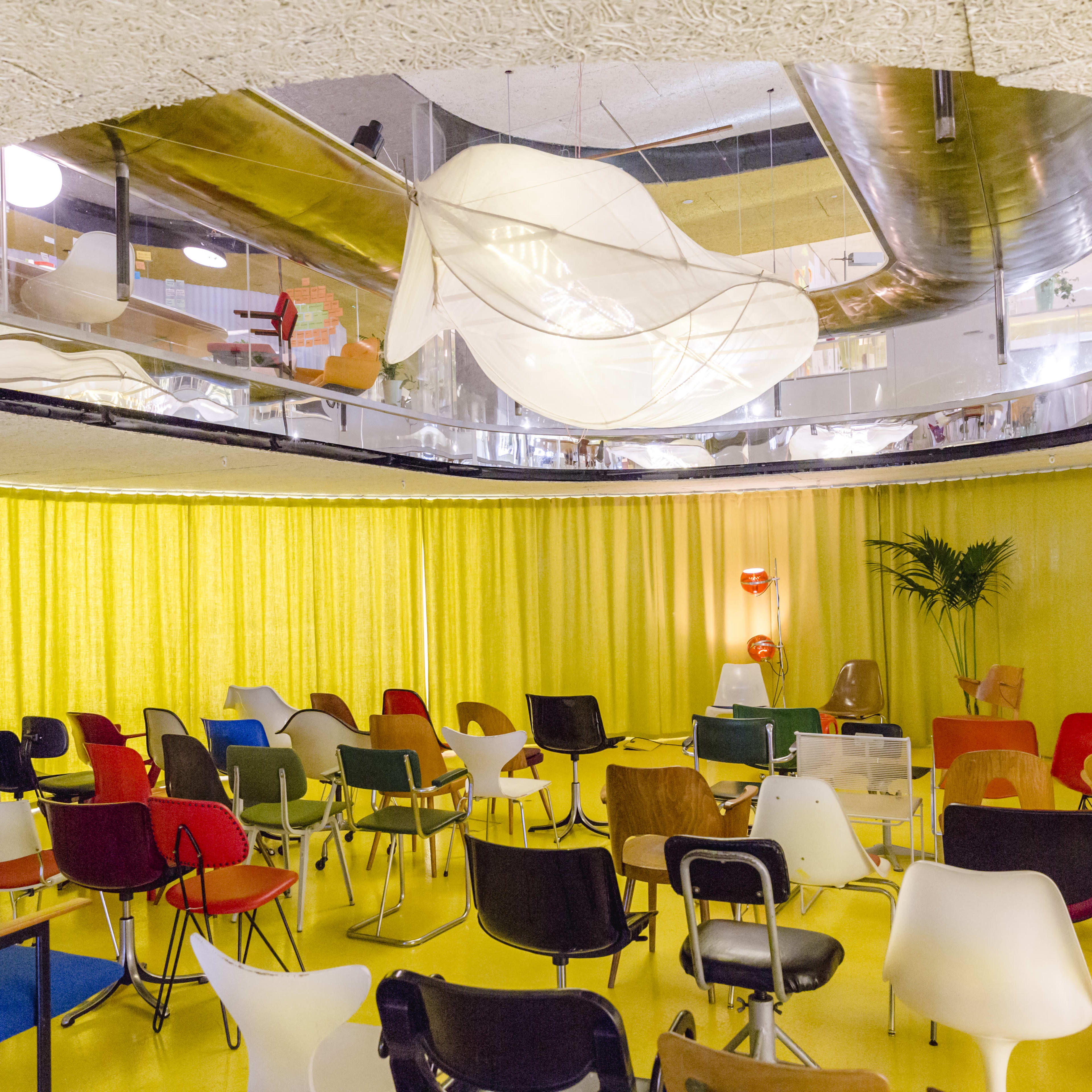 A brightly lit room features rows of colorful chairs arranged in a circular formation beneath a mirrored ceiling.