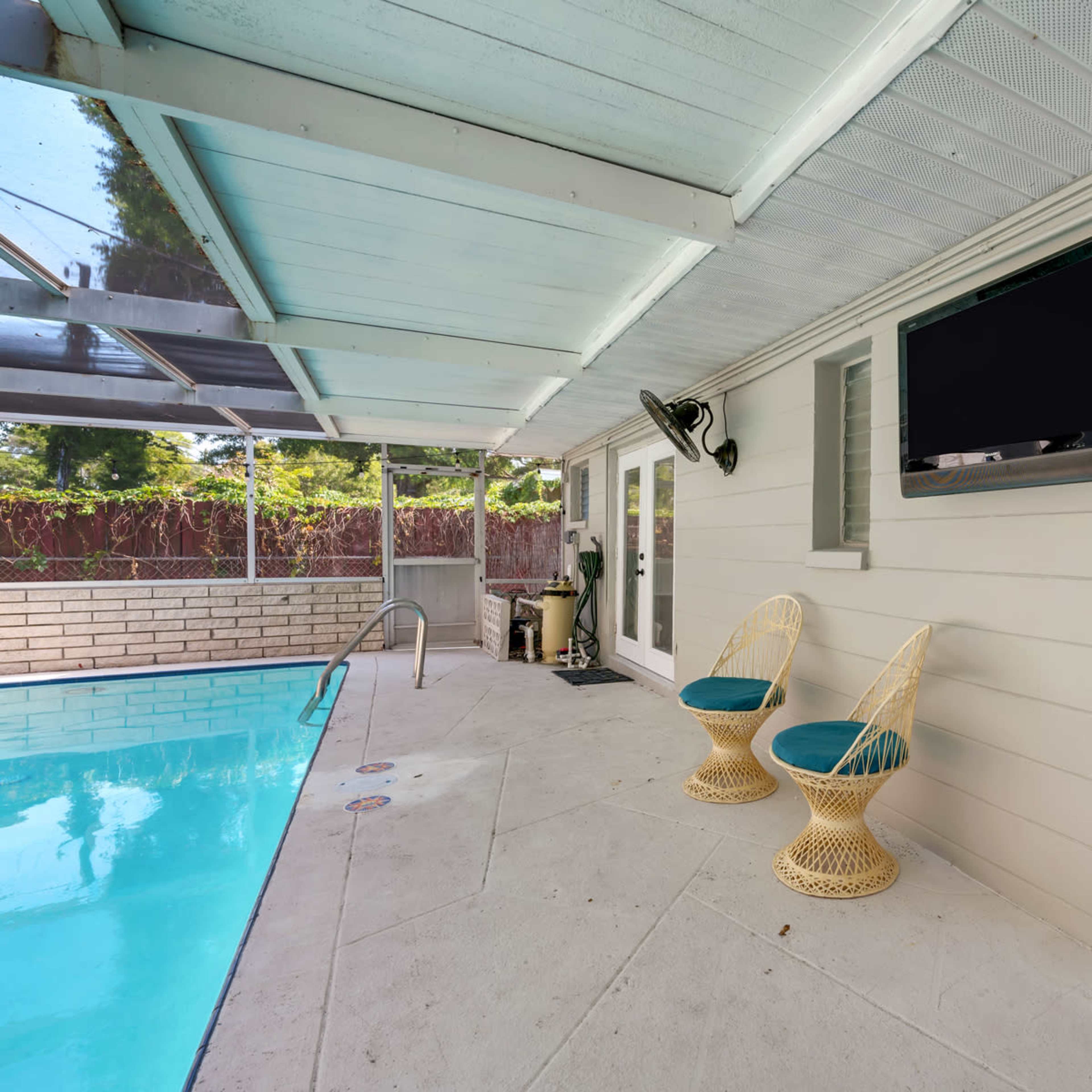 A covered patio area features two wicker chairs facing a swimming pool, with a television mounted on the wall nearby.