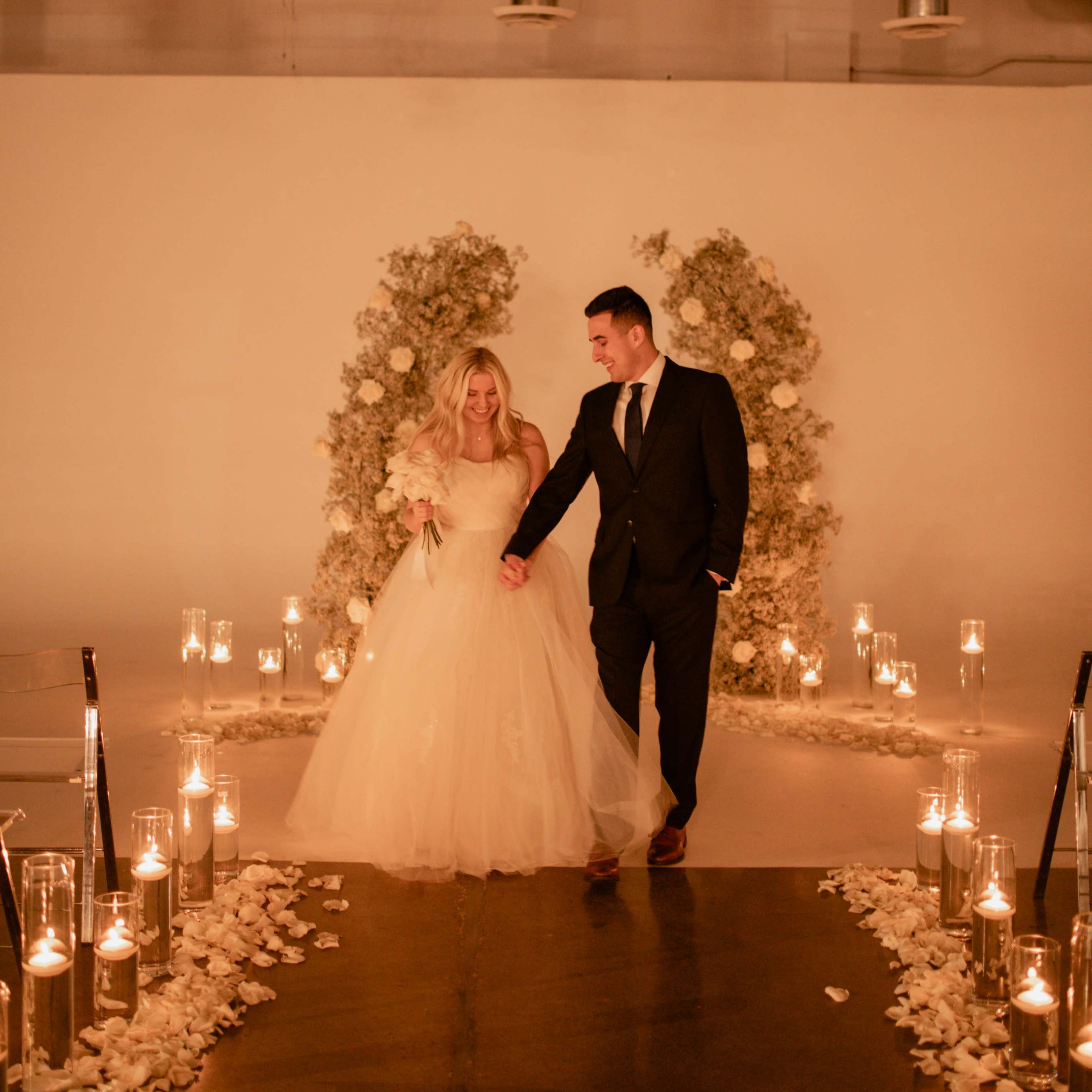 A bride and groom walk hand in hand down a candlelit aisle adorned with rose petals, flanked by floral arrangements.