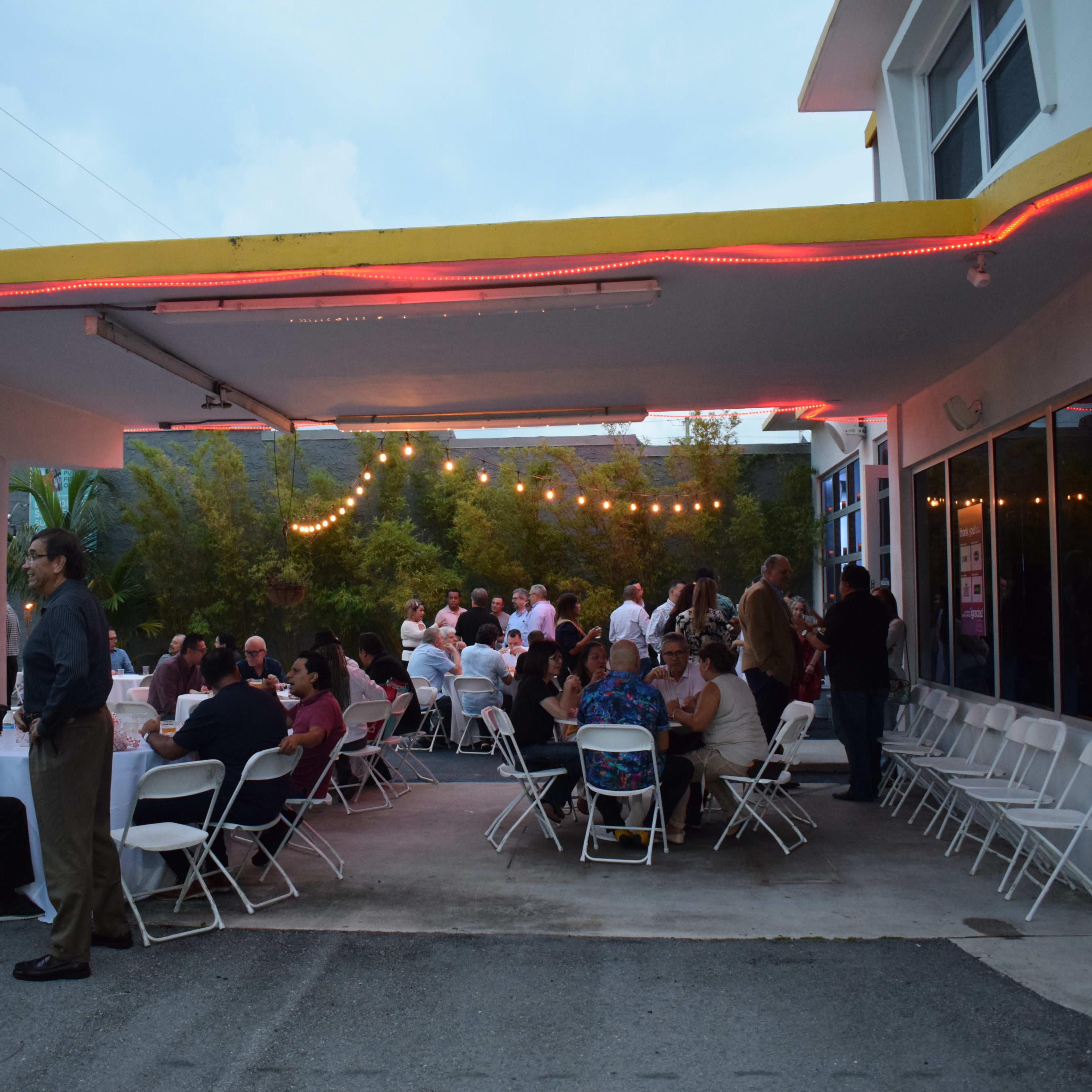 A group of people is gathering under a covered outdoor area, with tables set up for dining and string lights illuminating the scene.