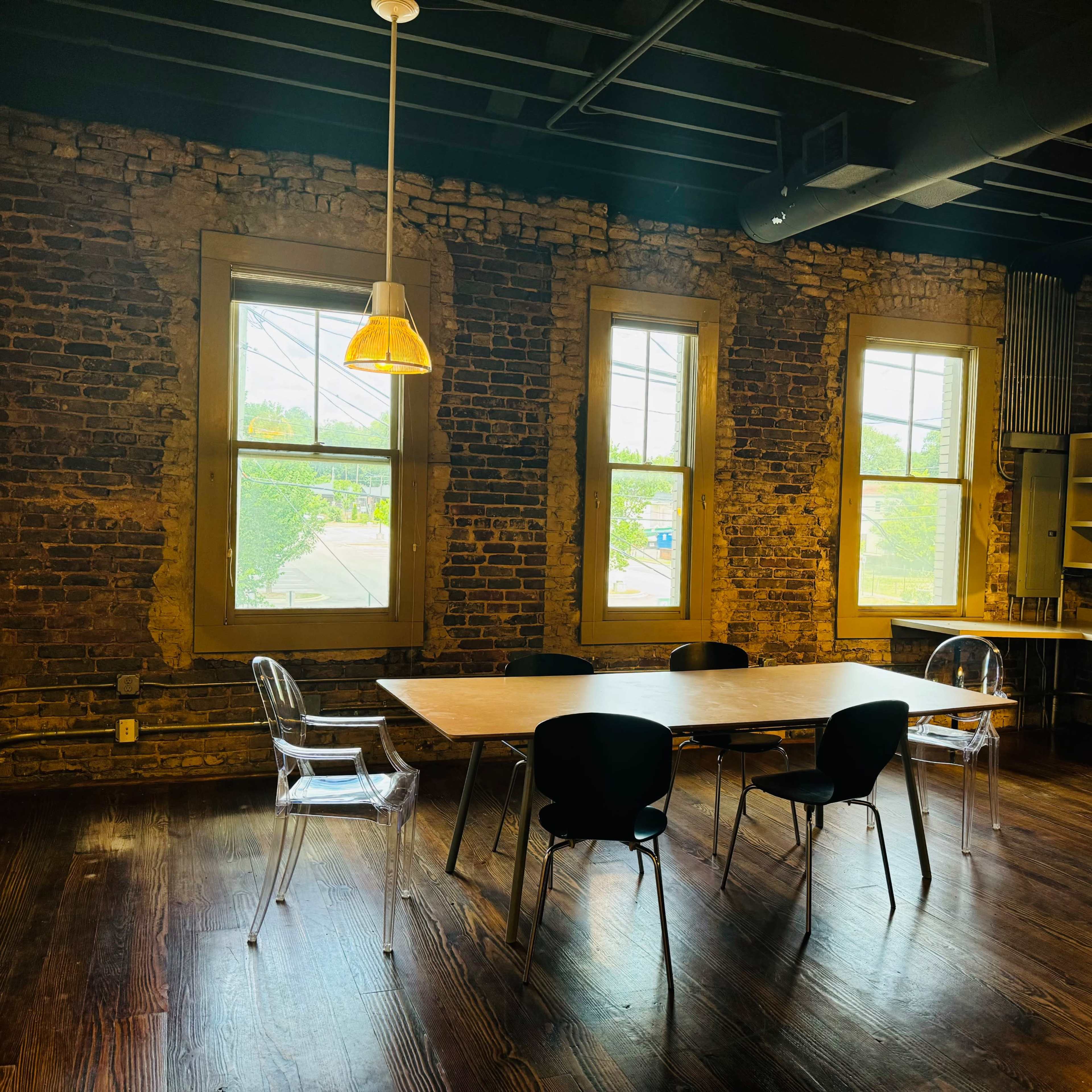 A workspace featuring a wooden table surrounded by black chairs, with exposed brick walls and large windows allowing natural light to enter.