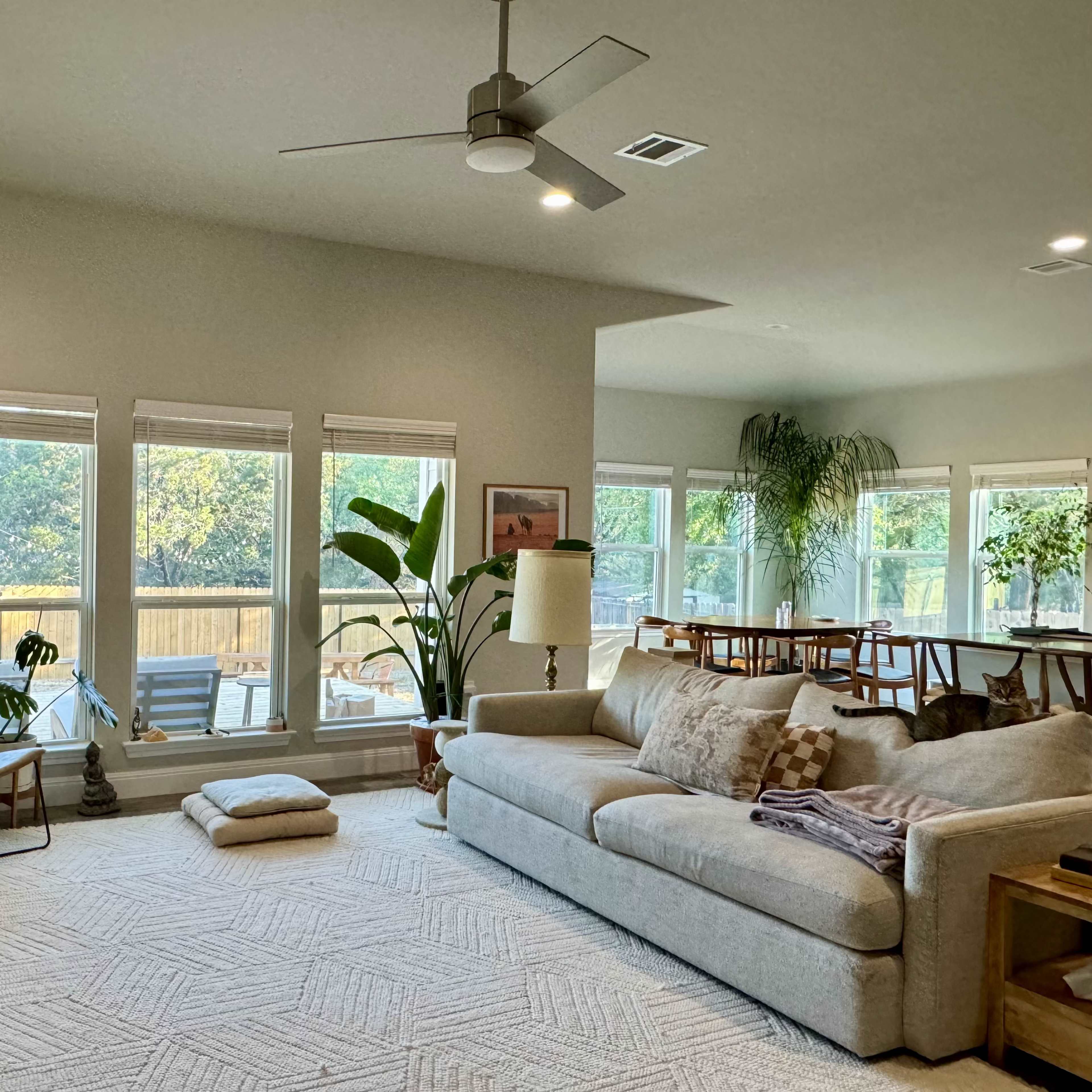 A spacious living room with large windows overlooking a patio, featuring a sectional sofa, a floor lamp, a rug, and a dining area in the background.