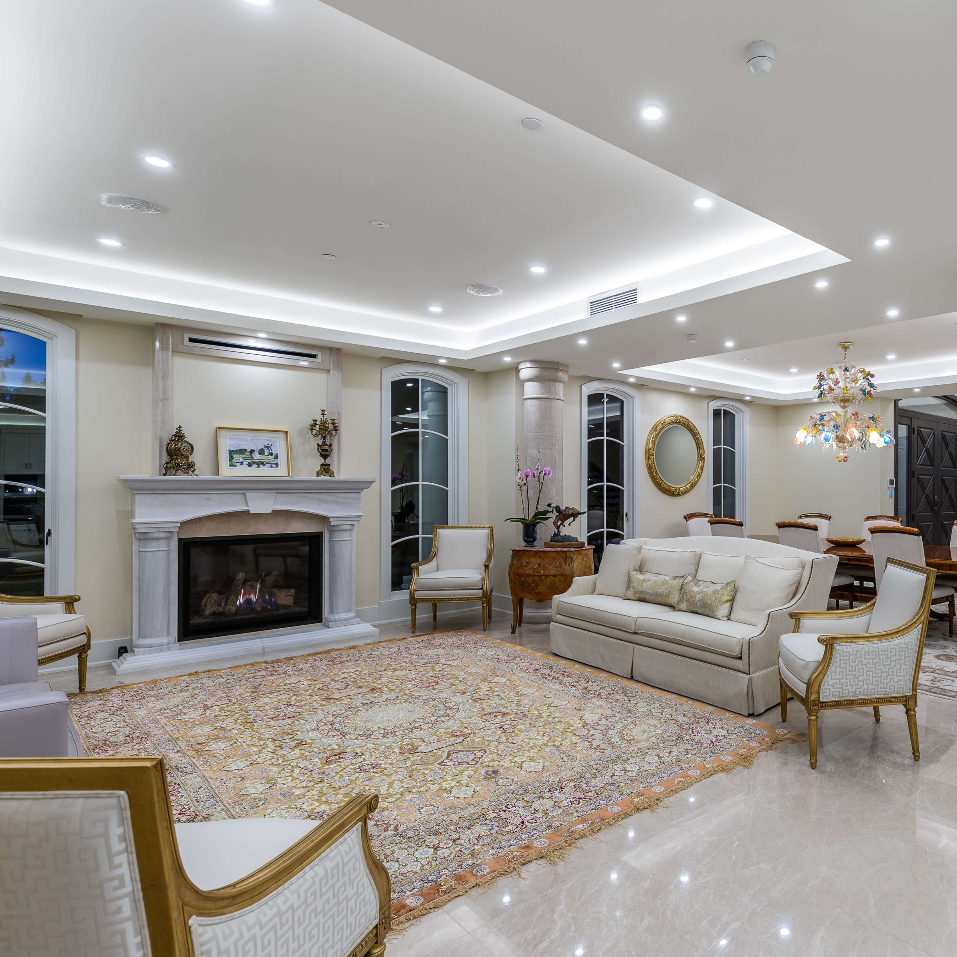 A luxurious living room featuring a fireplace, elegant seating, and a chandelier, with marble flooring and decorative pillars.