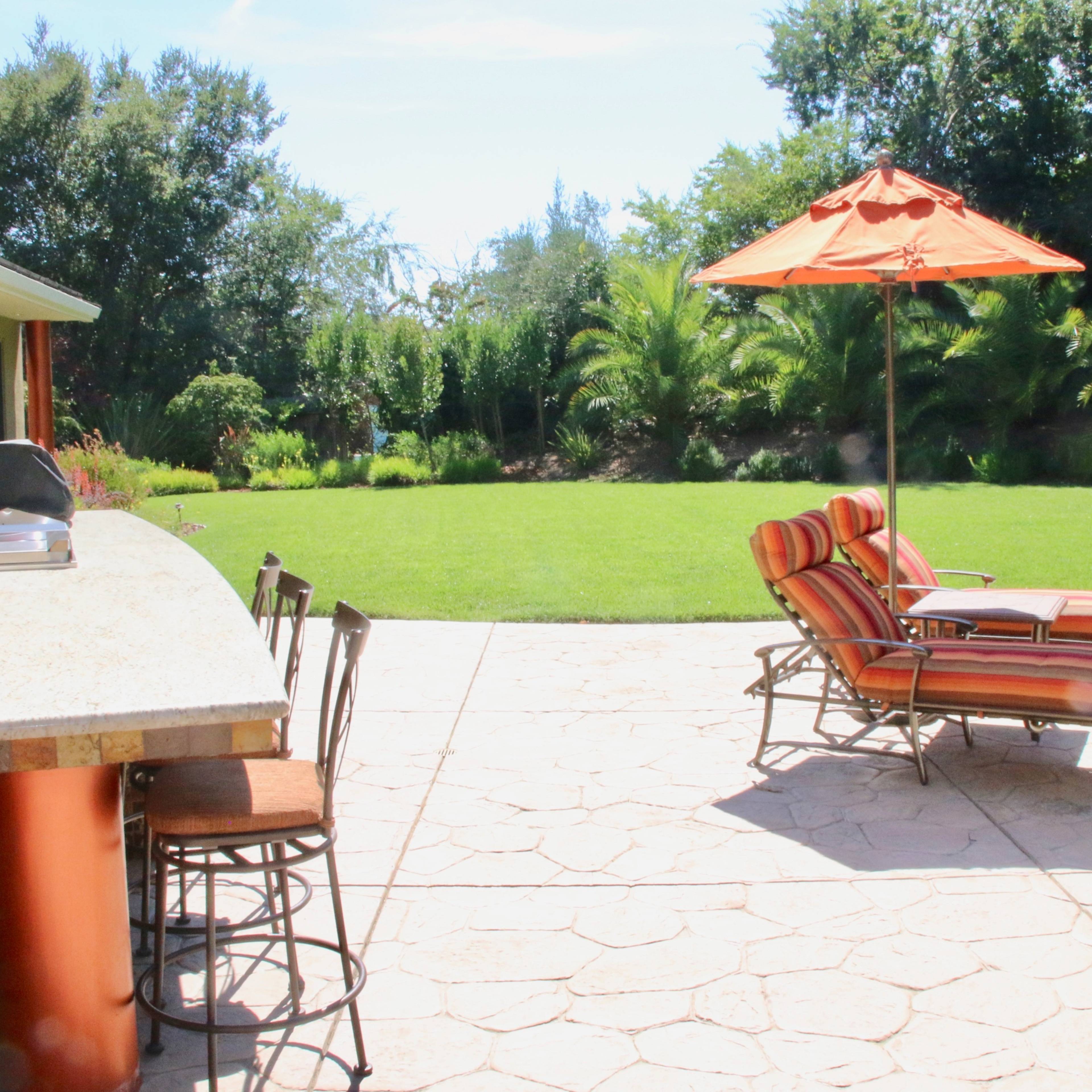 A patio area features a bar with stools, a lounge chair under an orange umbrella, and a green lawn surrounded by trees.