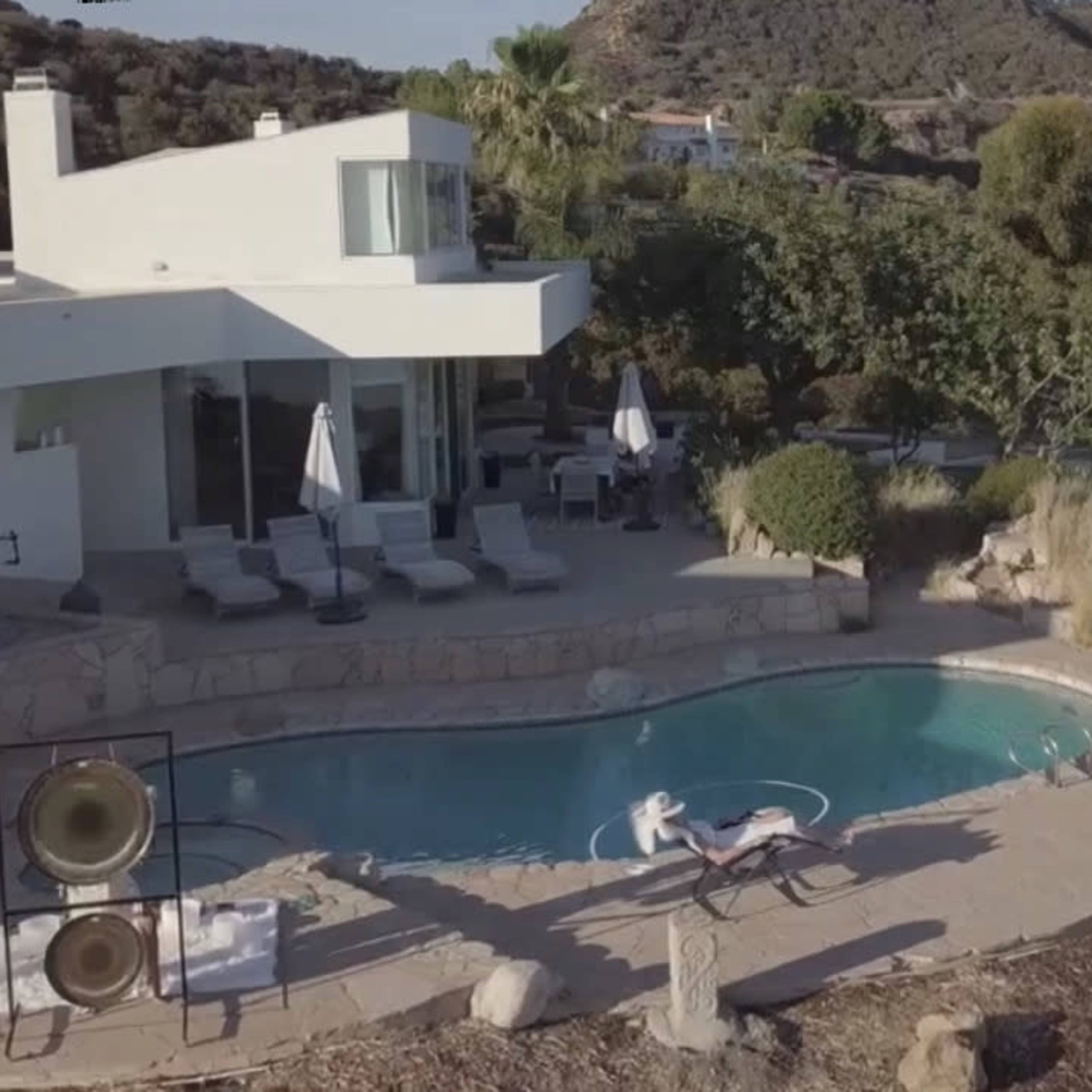 A modern house with large windows overlooks a swimming pool surrounded by lounge chairs and lush landscaping.